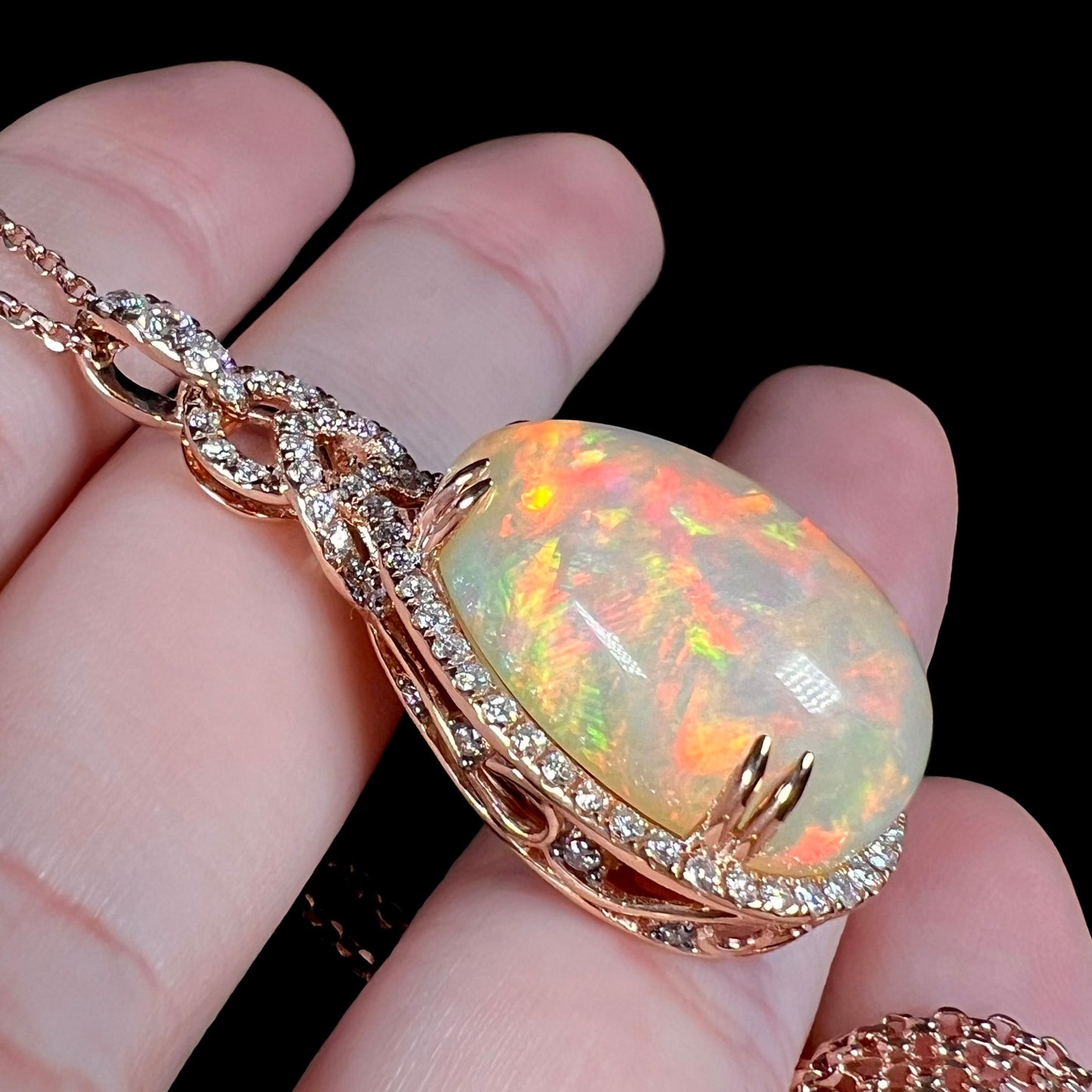Rose gold Ethiopian opal and diamond pendant shown resting on fingers from an angle against a black background.