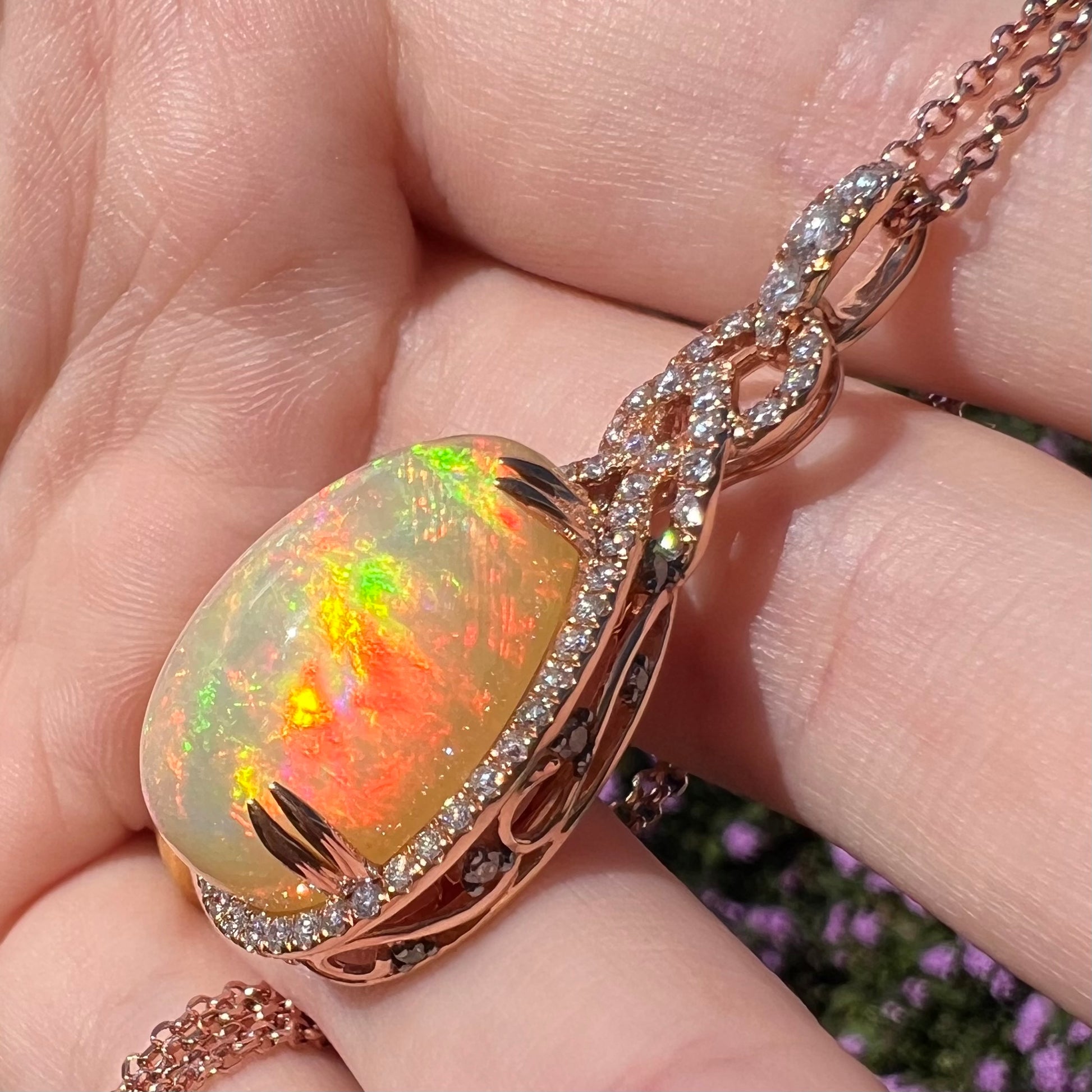 A rose gold Ethiopian opal and diamond pendant with vibrant red fire in the sunlight, shown from an angle held in a hand.