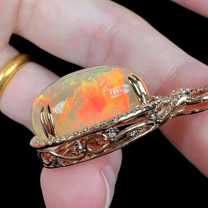 Hand holding a rose gold Ethiopian opal pendant, showing the pendant's filigree and diamond-accented side basket.  The opal has a bright red flash of color.