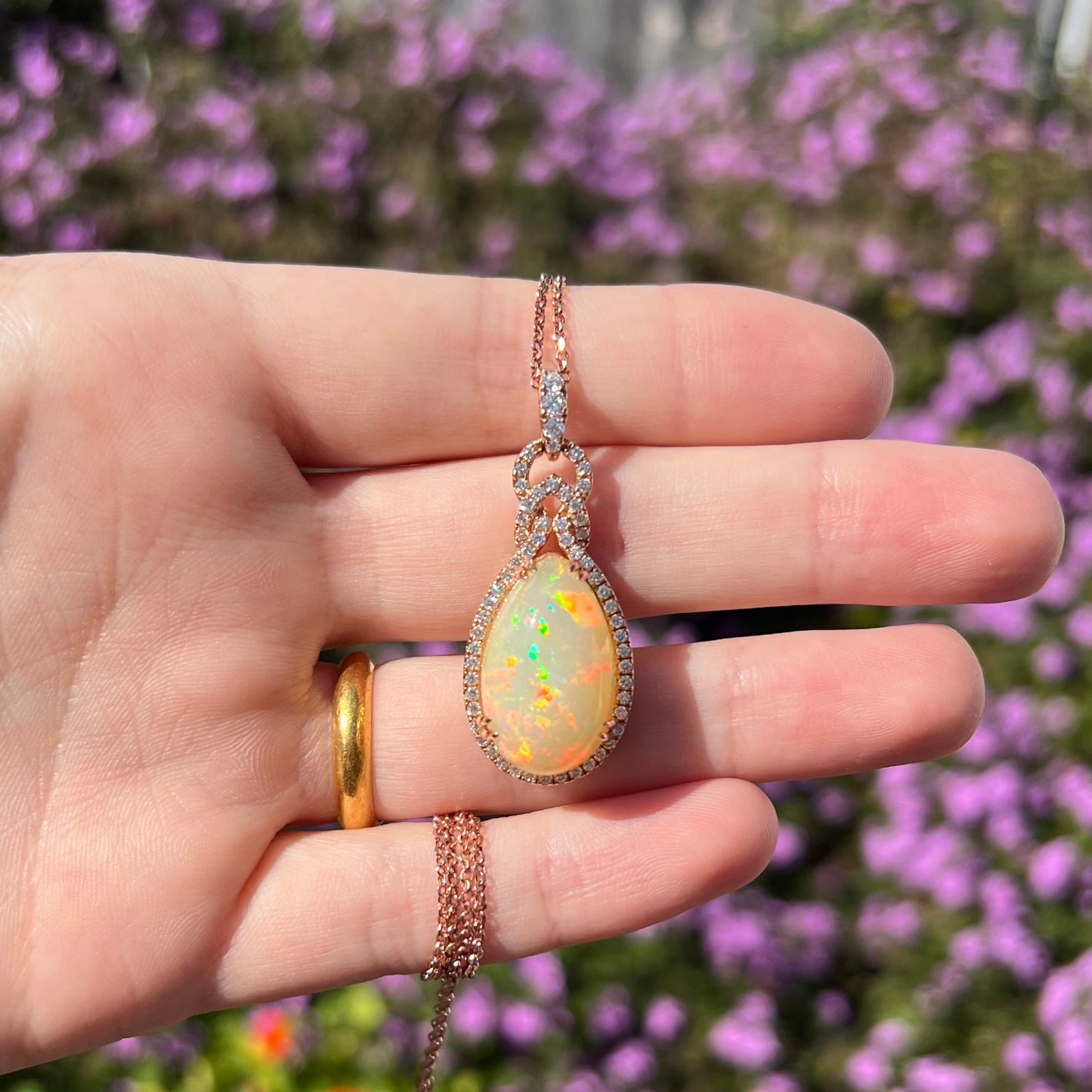 A teardrop shaped opal and diamond halo necklace in rose gold, shown held in a hand under natural sunlight against a blurred outdoor background.