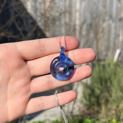 A round blue glass pendant with a suspended red lab-created opal chip inside, shown held in a hand in the sunlight against an outdoor background.