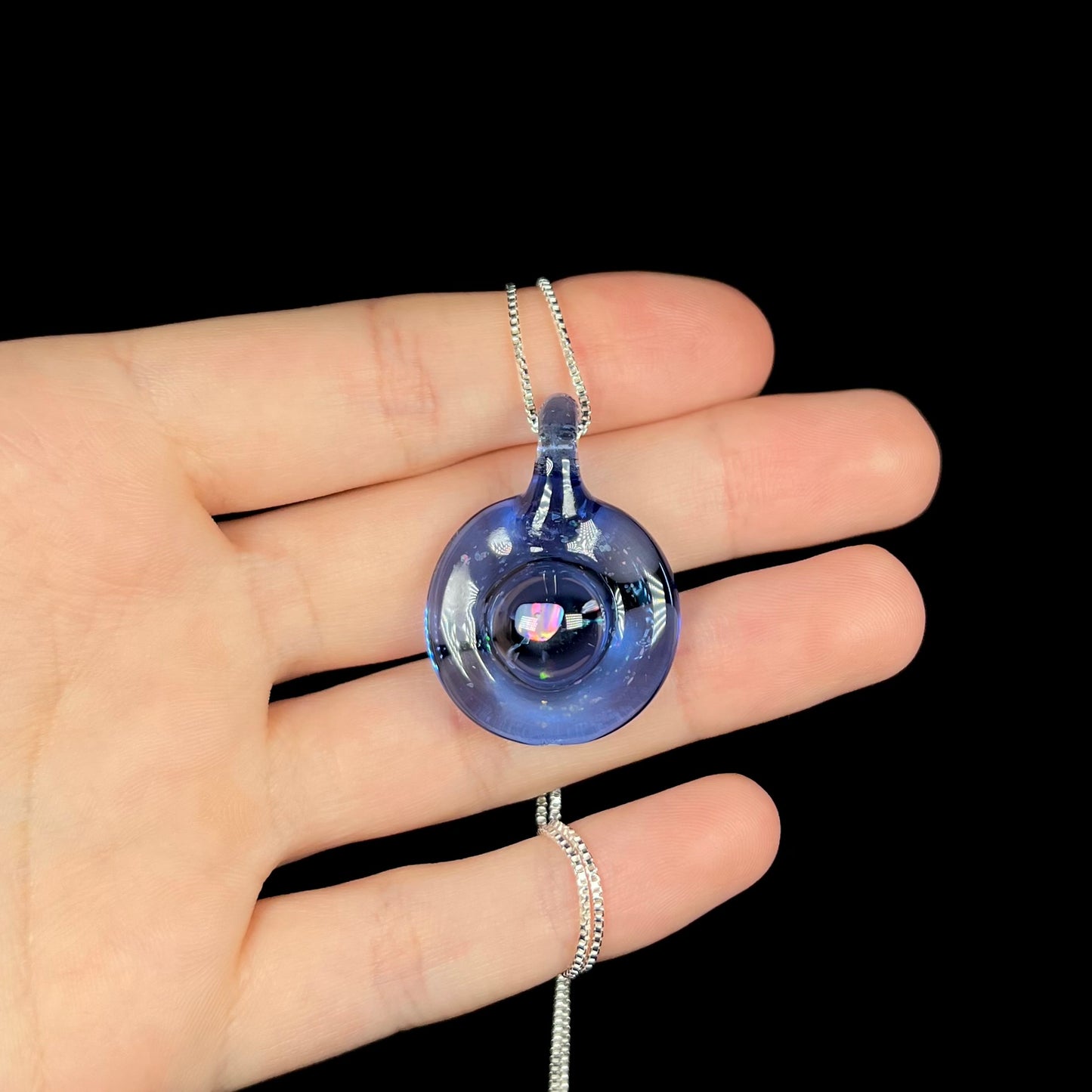 A hand holding a round, blue glass opal necklace against a black background.  The silver chain wraps around the pinky finger.