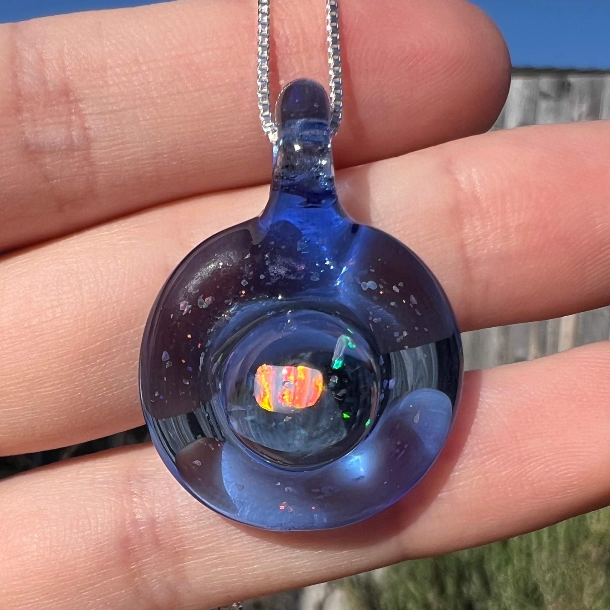 Close-up of an opal in a round, blue glass pendant shown close-up in a hand in the sunlight.  The opal has red color play.