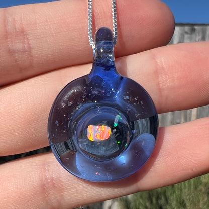 Close-up of an opal in a round, blue glass pendant shown close-up in a hand in the sunlight.  The opal has red color play.