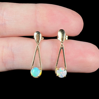 Pair of round cut opal dangle earrings in yellow gold shown close-up between two fingers against black backdrop.