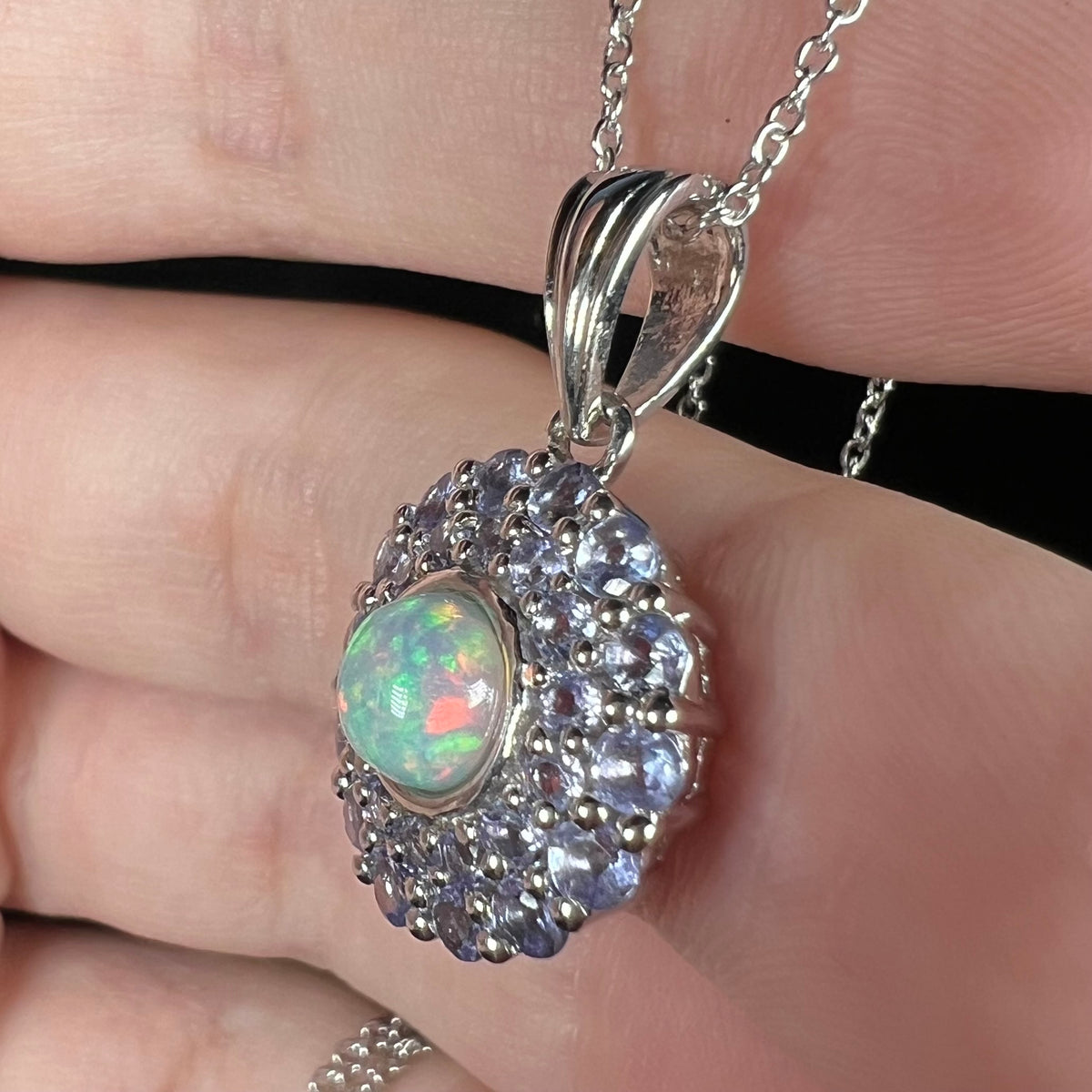 Angled side close-up of a round Ethiopian fire opal pendant in sterling silver with a tanzanite halo, held in a hand under LED studio lights.
