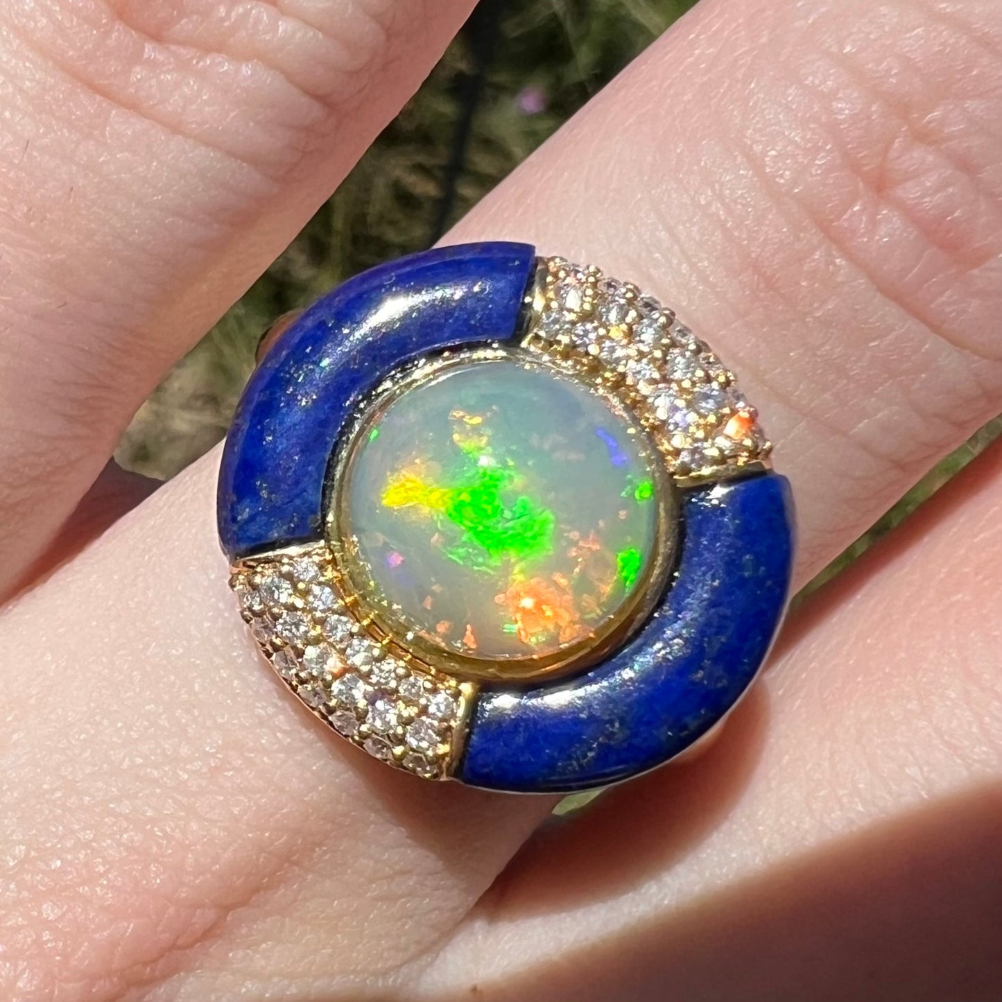A close-up of a gold ring worn on a finger, featuring a round iridescent Ethiopian opal center surrounded by a halo of blue lapis lazuli and small clear pavé diamonds in natural light.