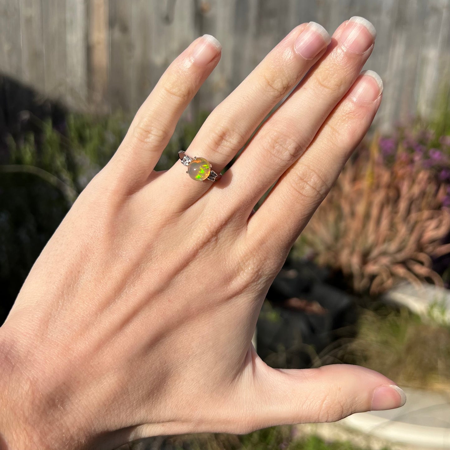 A platinum fire opal and diamond engagement ring on a hand in the sunlight, showcasing the round opal's green play-of-color.