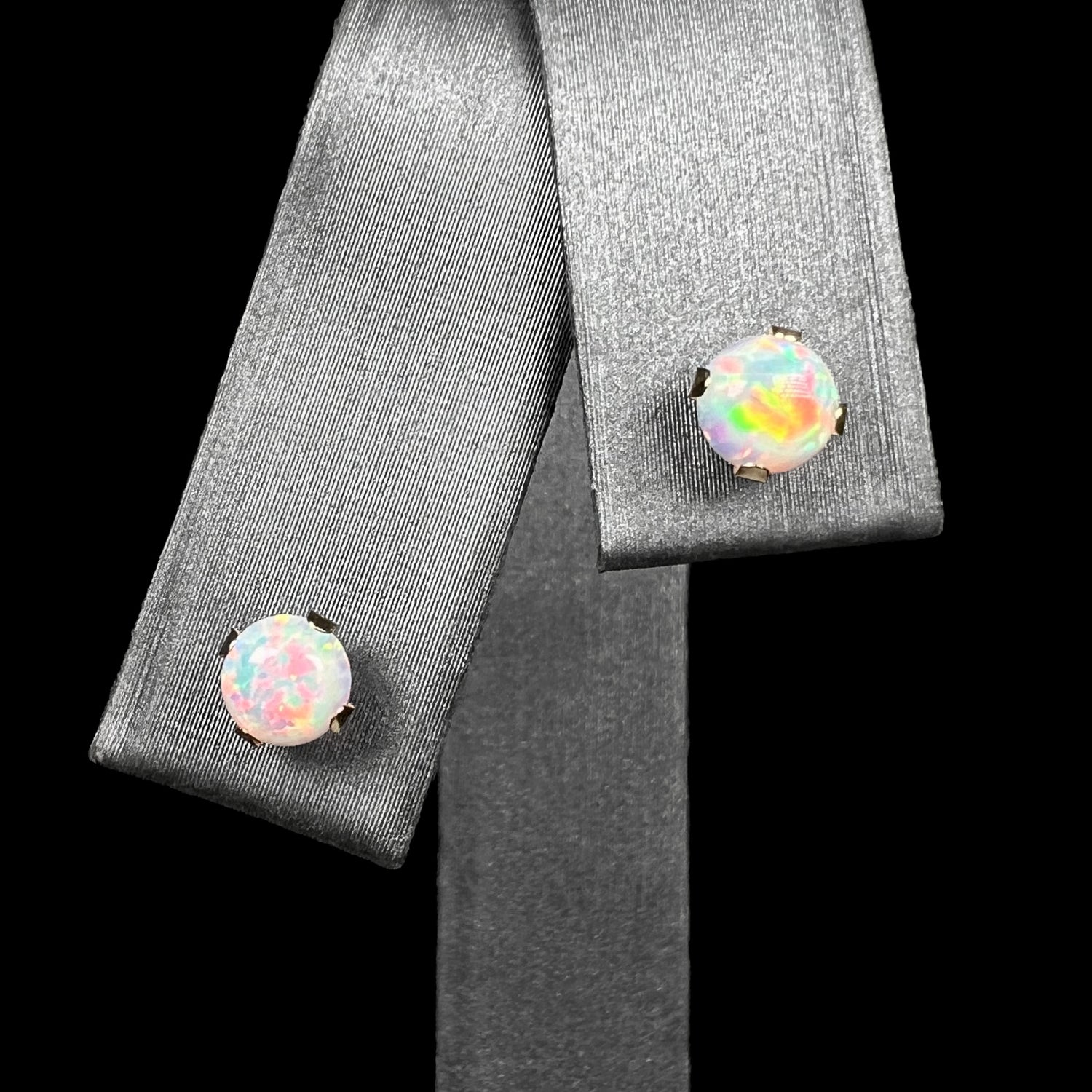 A pair of women's dainty round lab-created opal stud earrings prong-set in 14k yellow gold, shown from front close-up view on earring display against black background.