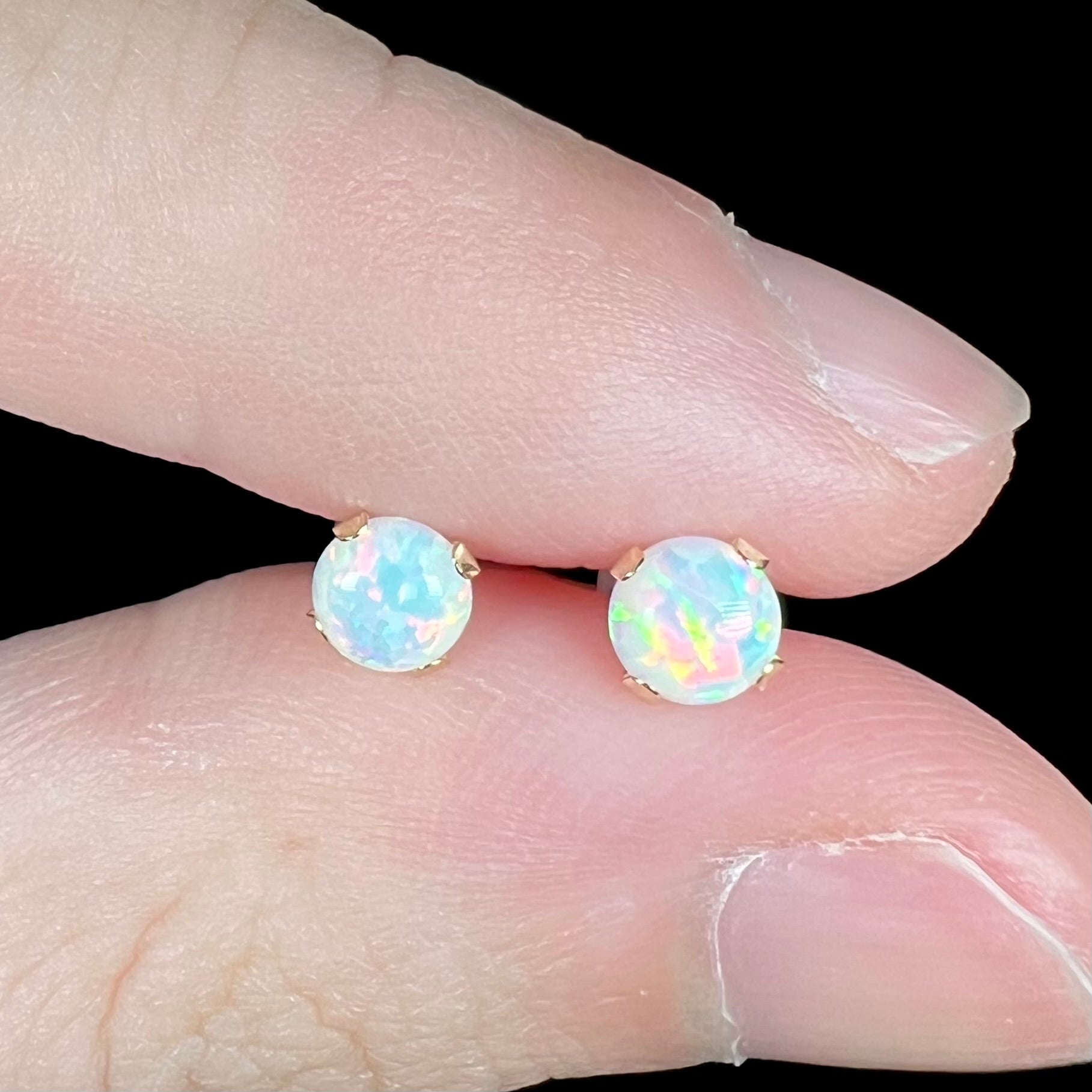 Close-up macro view of round lab-created opal stud earrings in 14k yellow gold settings, held between two fingers under cool LED studio light.