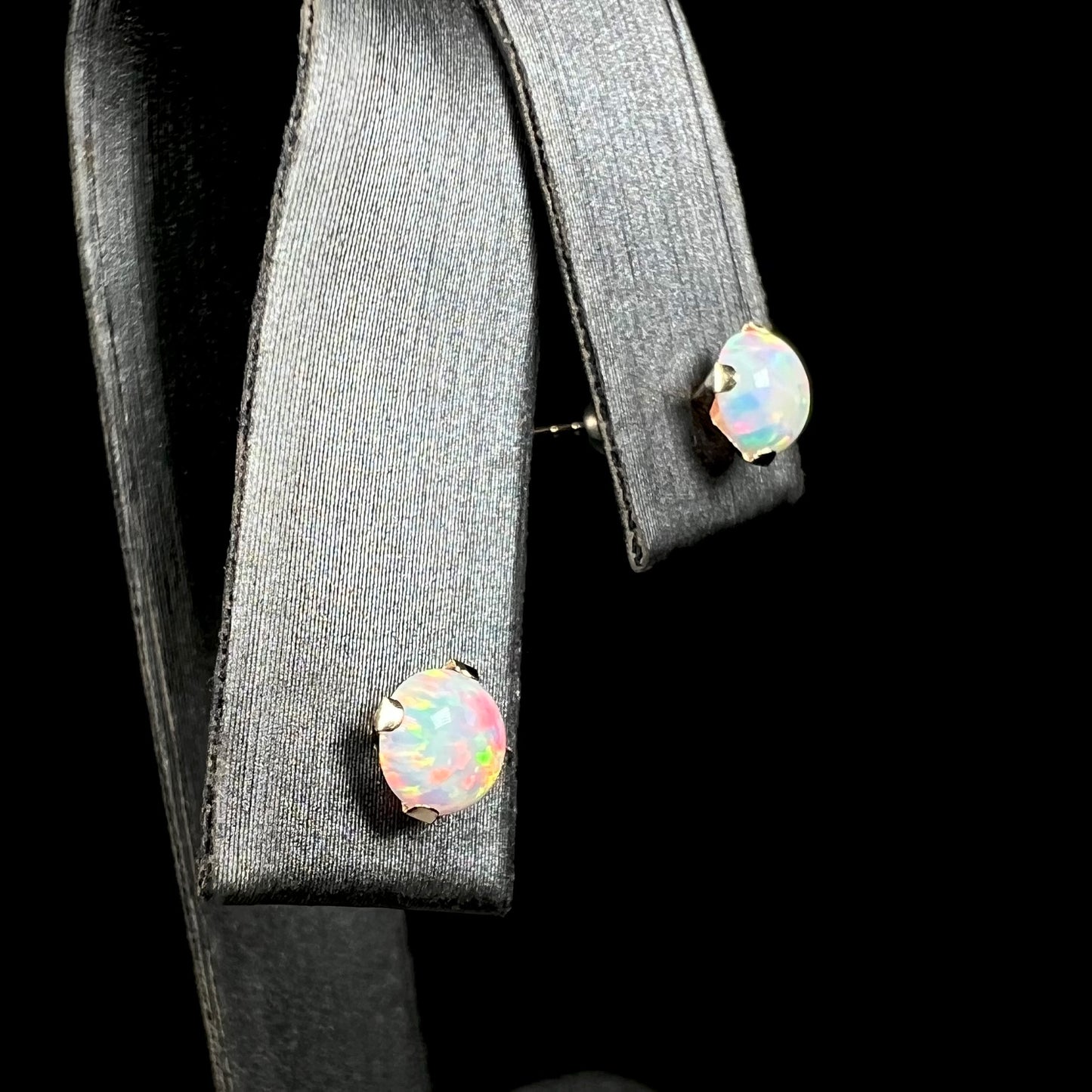 A pair of round lab-created opal stud earrings in 14k yellow gold, shown close-up from angle view in earring display.  The closest opal has red flashes of color.
