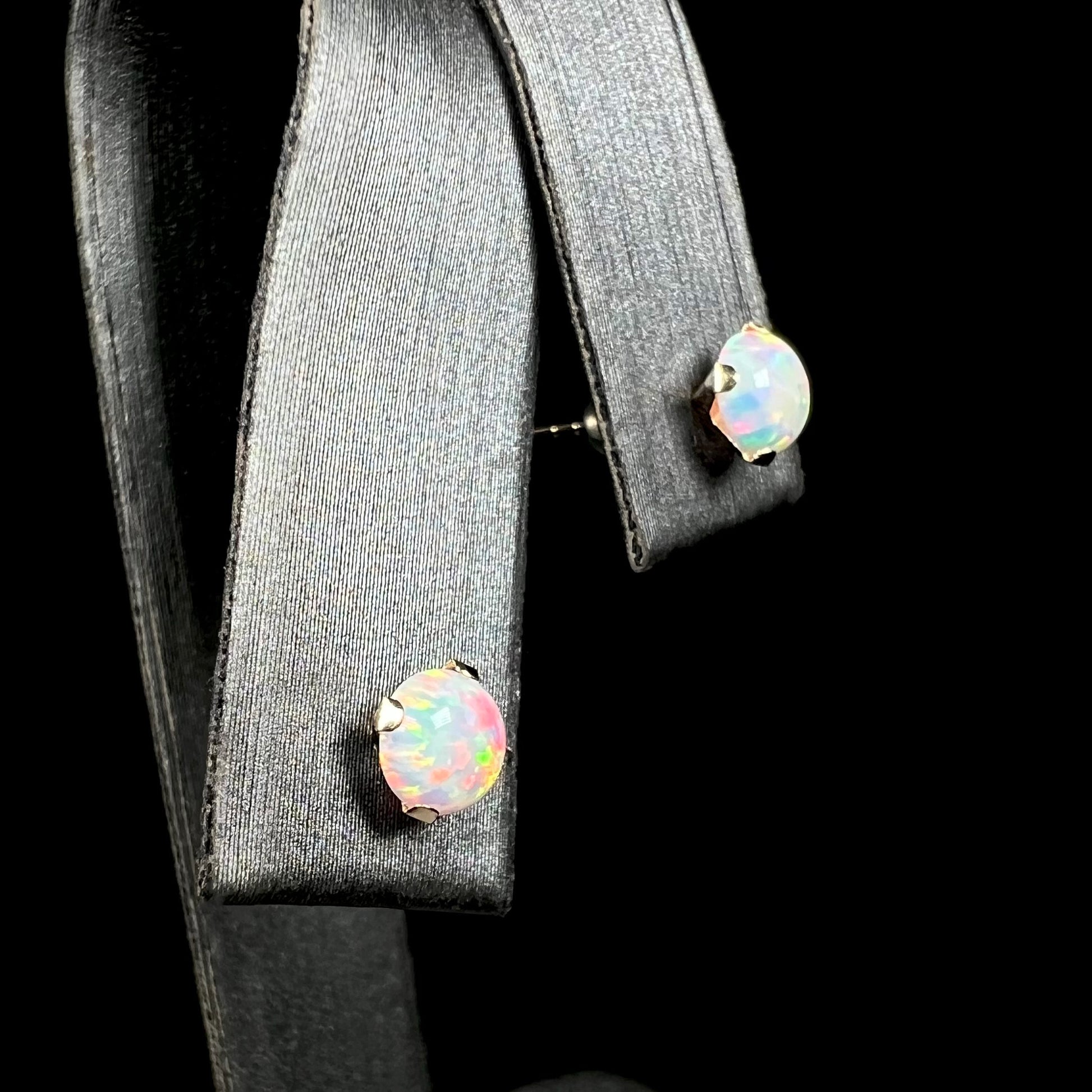 A pair of round lab-created opal stud earrings in 14k yellow gold, shown close-up from angle view in earring display.  The closest opal has red flashes of color.