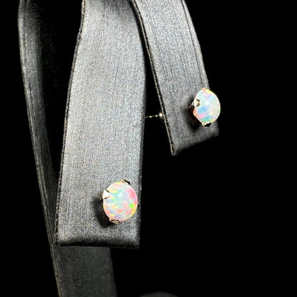 A pair of round lab-created opal stud earrings in 14k yellow gold, shown close-up from angle view in earring display.  The closest opal has red flashes of color.
