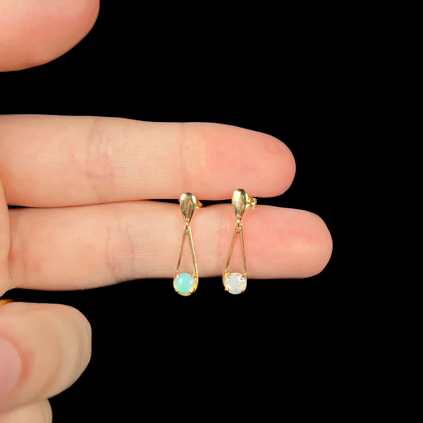 Round opal drop earrings in yellow gold held between two fingers against black background under studio light.