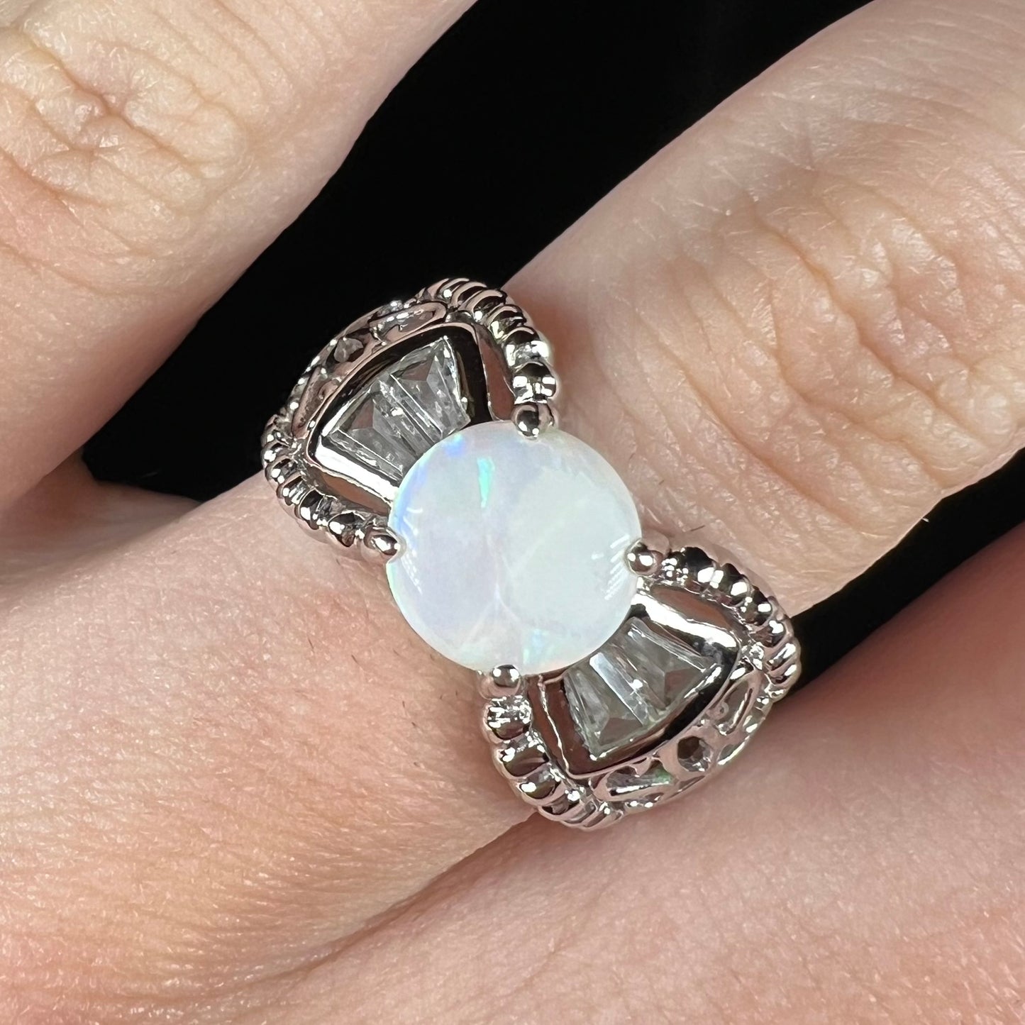 Front view of a round Australian white opal and CZ ring in white base metal worn on a finger.