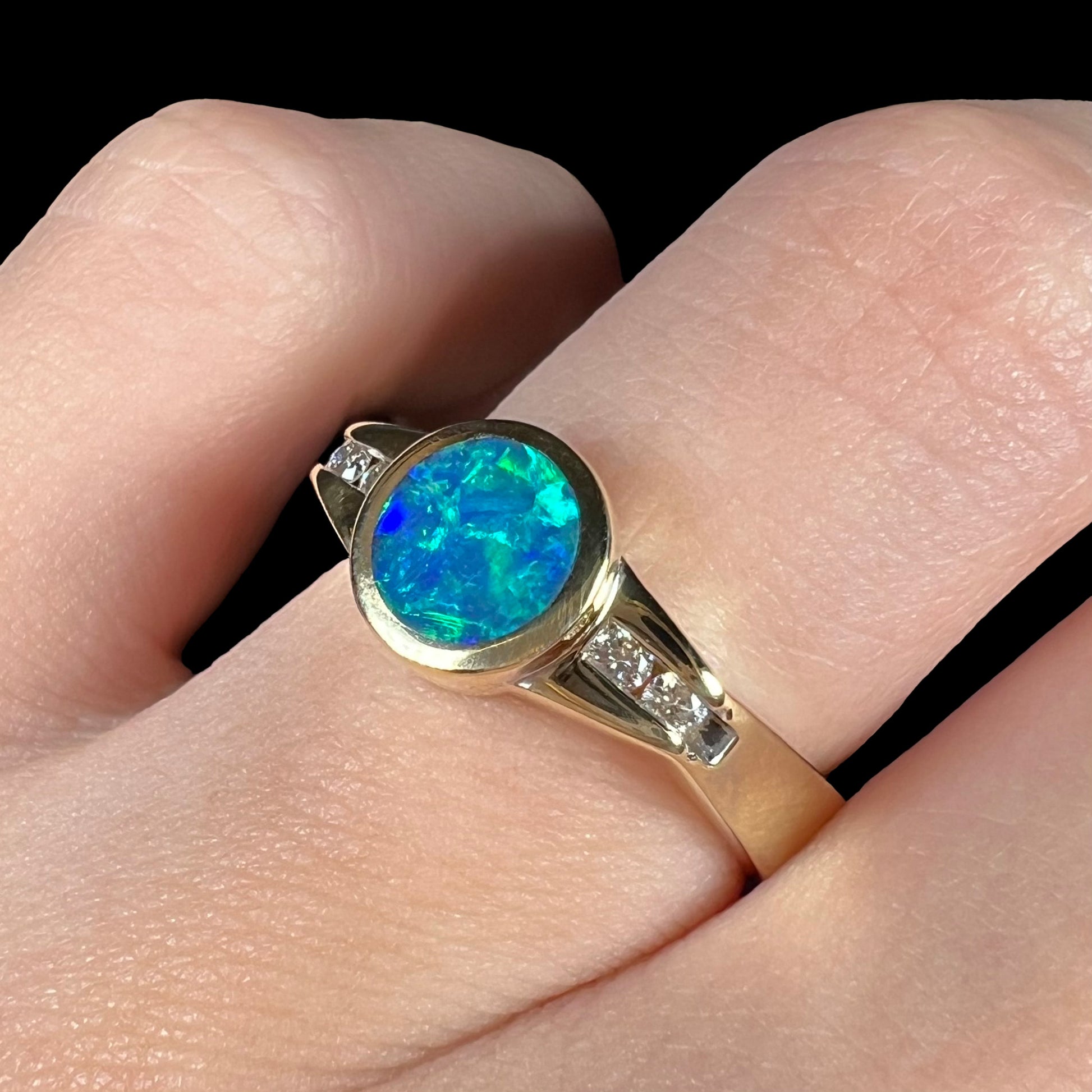 Macro view of a round Queensland boulder opal ring with diamond accents and blue color play, shown from an angle on a finger.