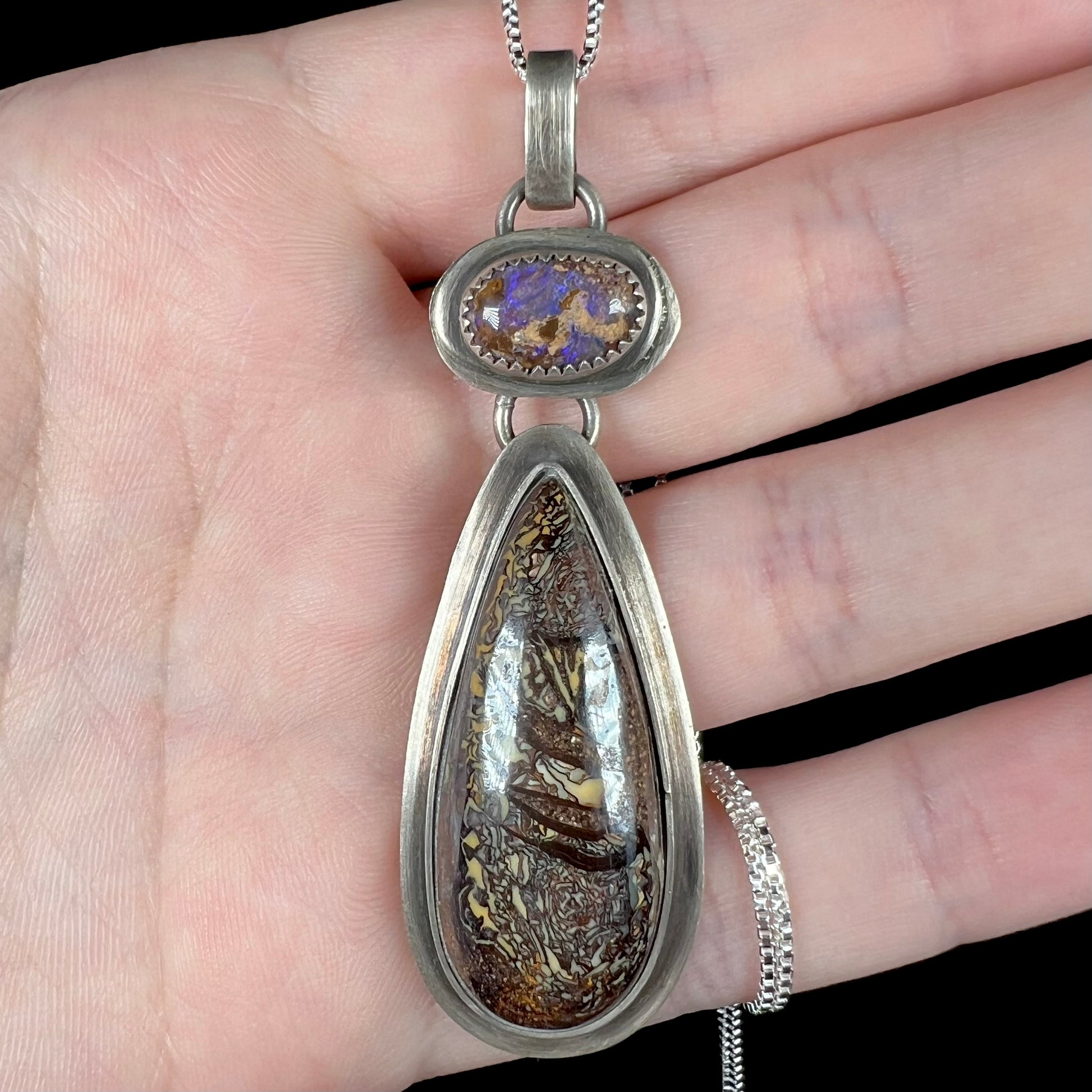 Close-up of a rustic sterling silver boulder opal pendant held in a hand under cool LED lighting.