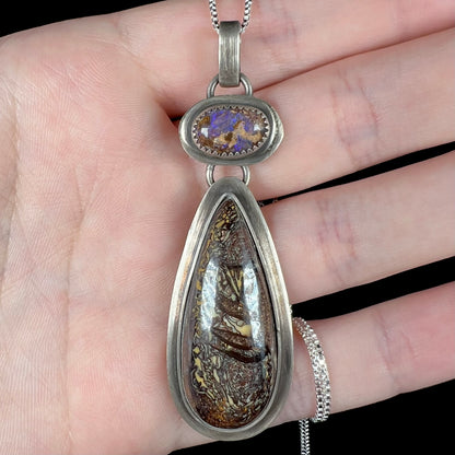 Close-up of a rustic sterling silver boulder opal pendant held in a hand under cool LED lighting.