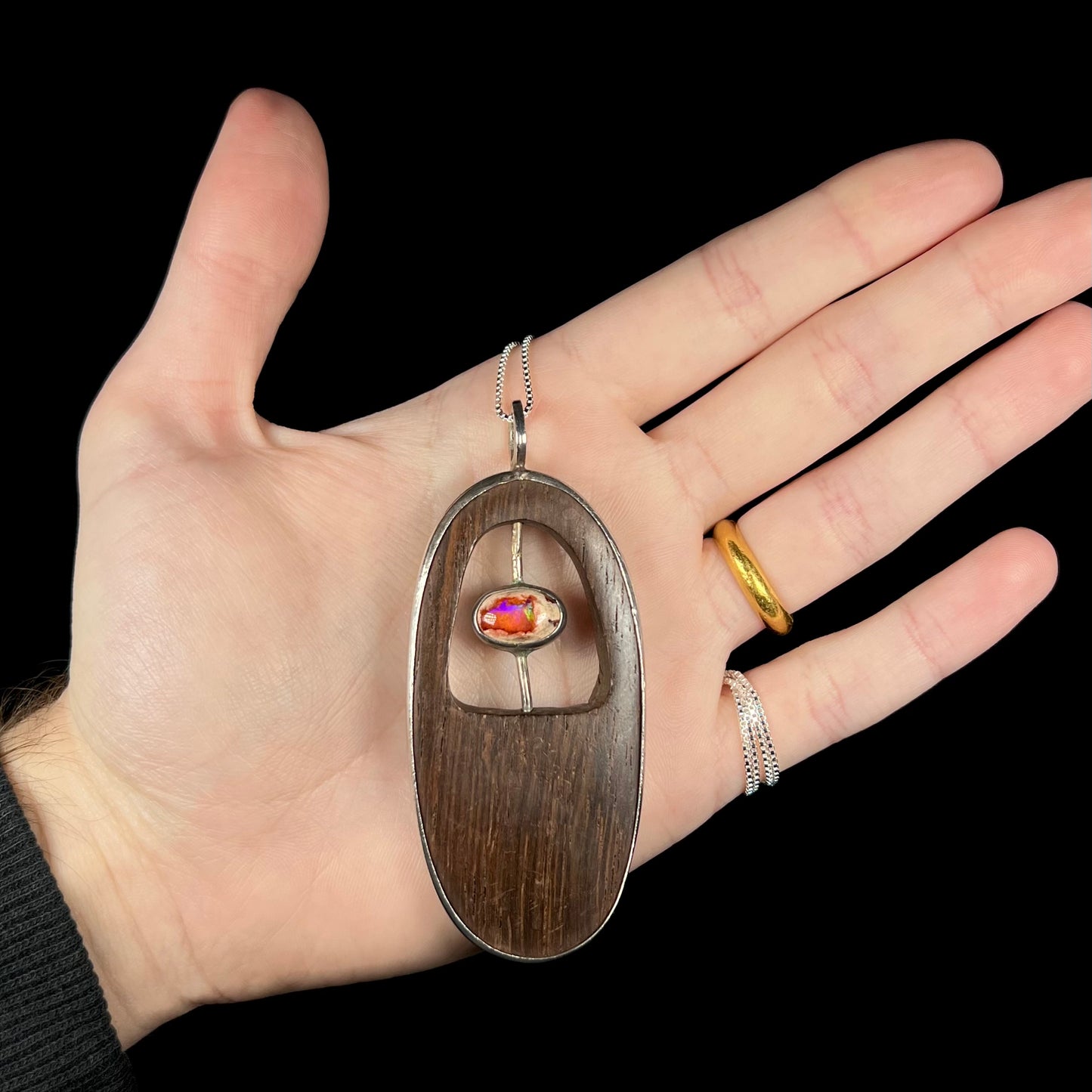 Hand holding a large, oval-shaped rustic Mexican fire opal and carved wood men's necklace against a black background.
