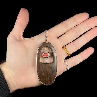 Hand holding a large, oval-shaped rustic Mexican fire opal and carved wood men's necklace against a black background.