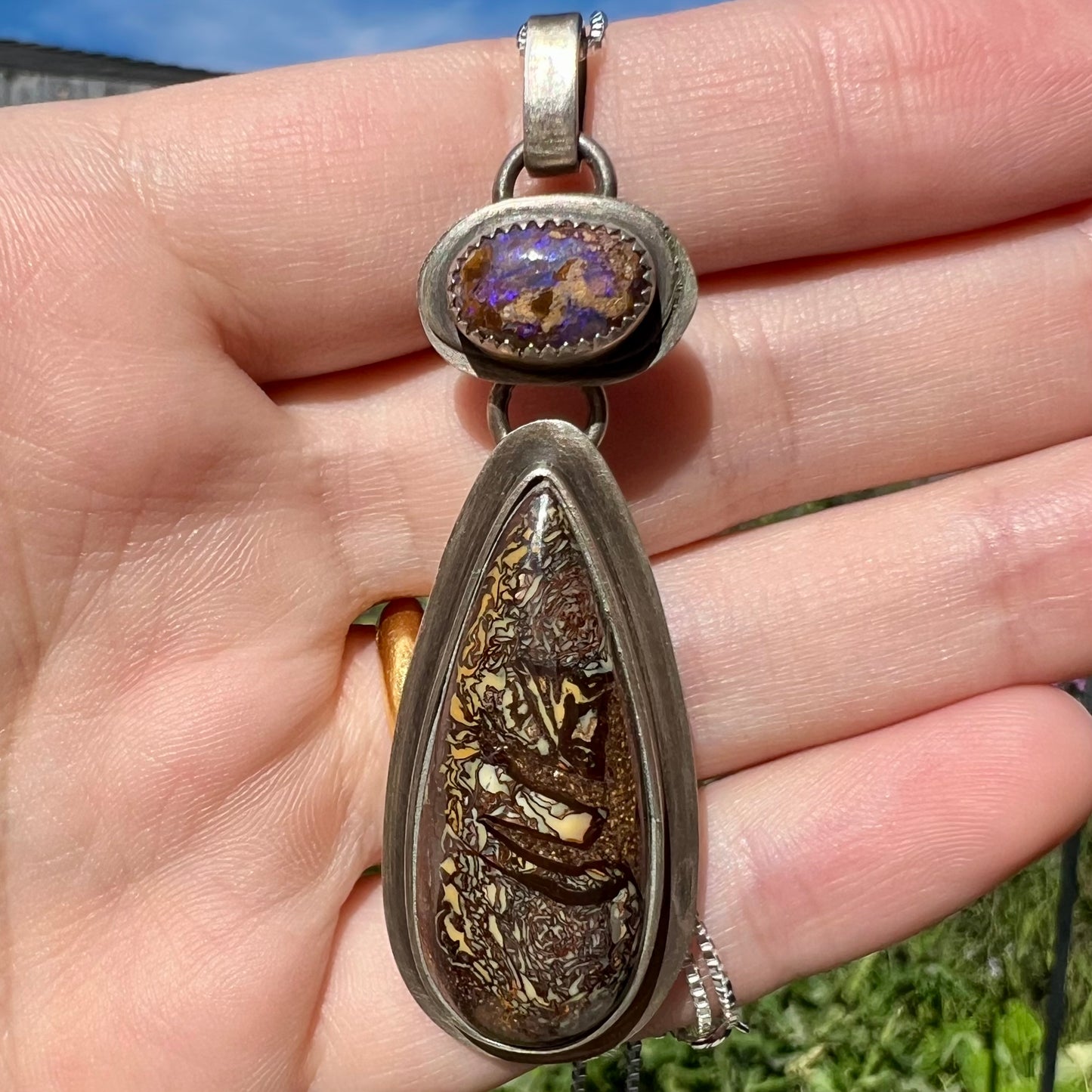 Rustic silver boulder opal pendant held in a hand against a natural background in daylight.