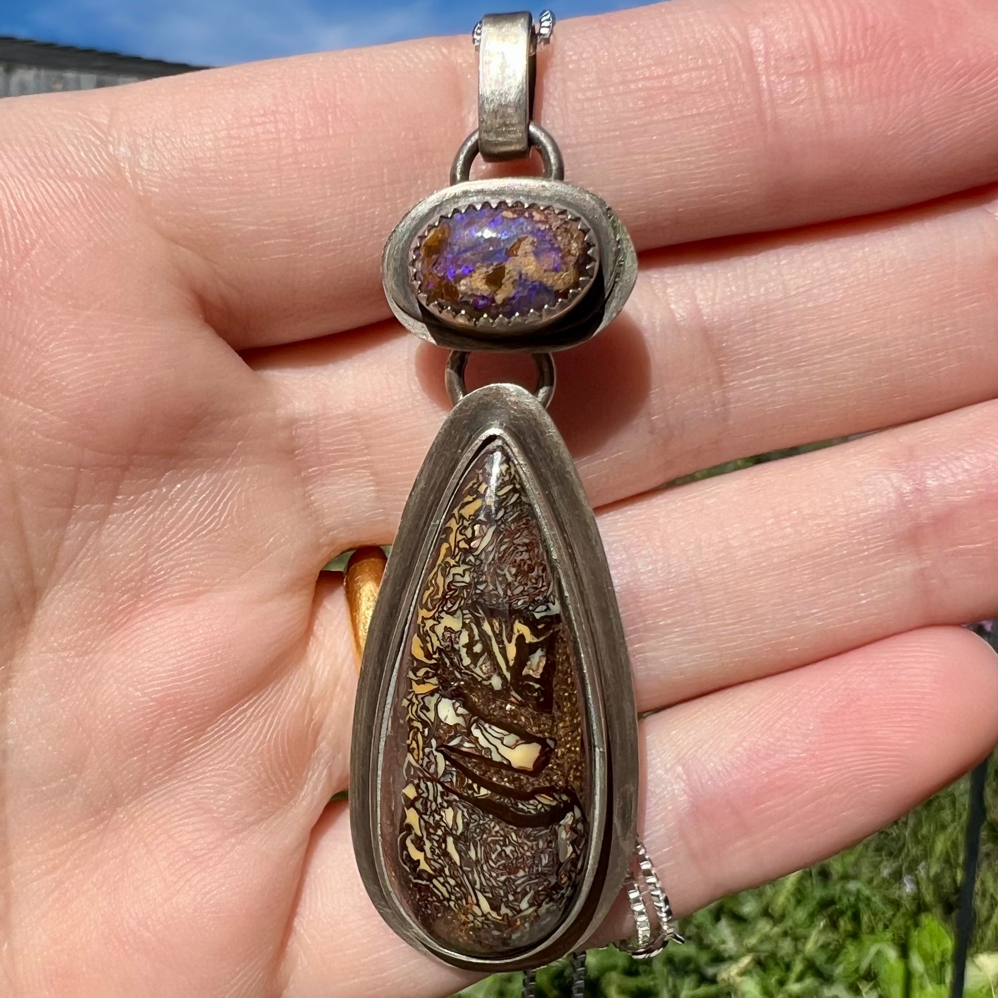 Rustic silver boulder opal pendant held in a hand against a natural background in daylight.