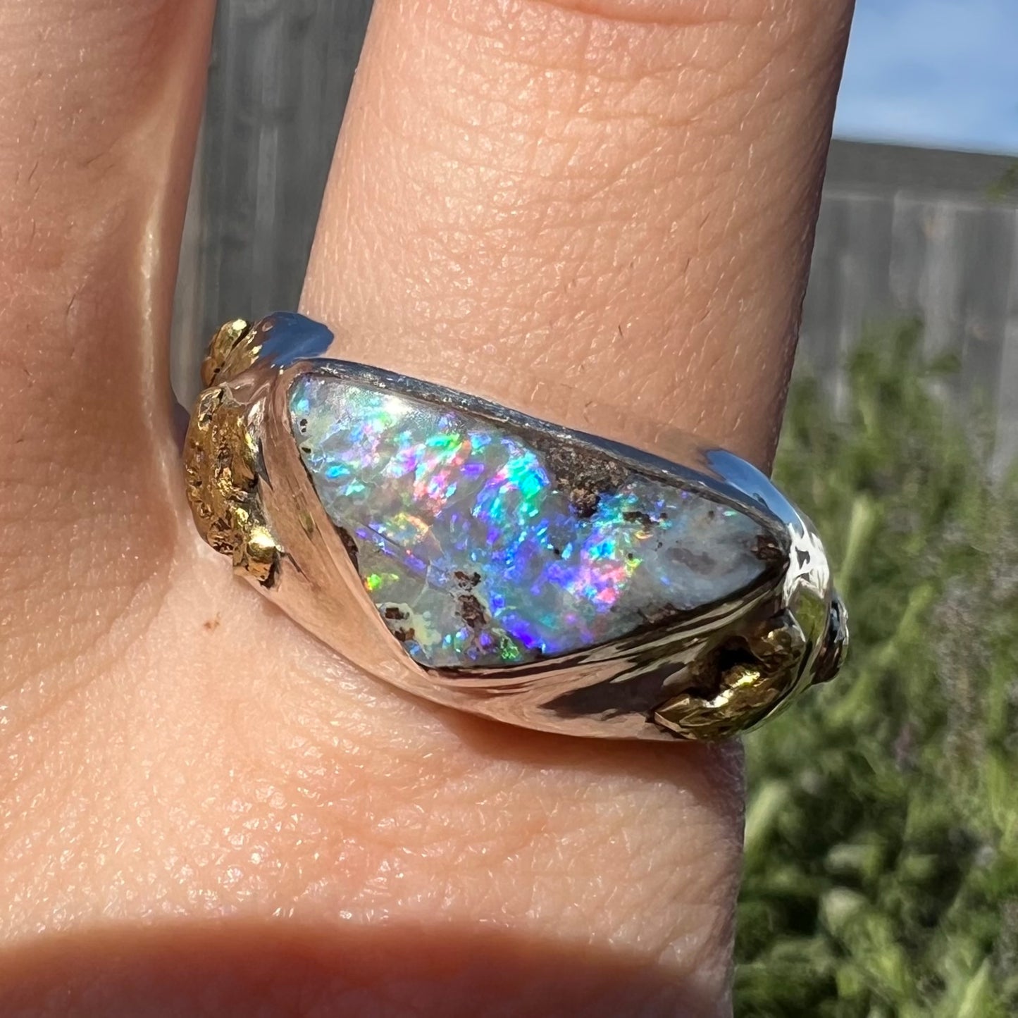 Close-up of a rustic-style men's sterling silver boulder opal ring worn on a finger in sunlight.  The triangular opal has blue and magenta fire.