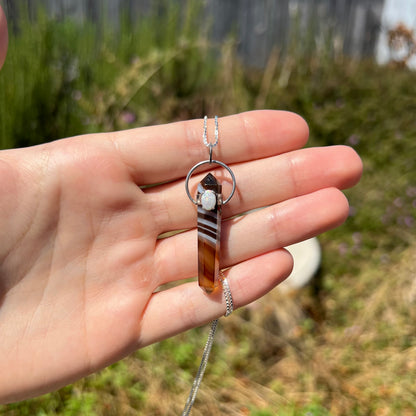 A sardonyx and opal pendant made from silver, held in the sunlight by a hand.