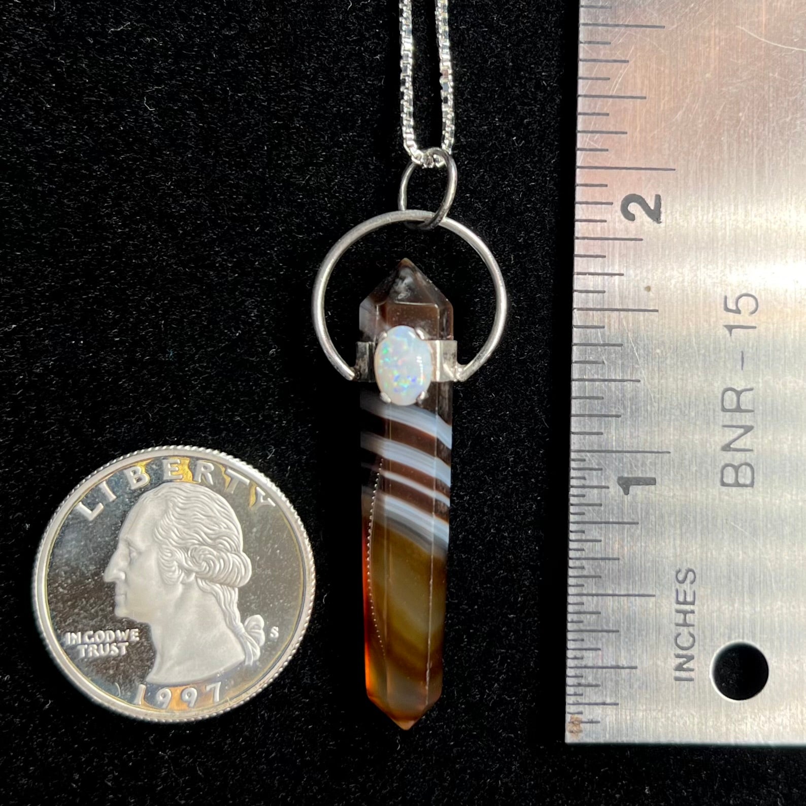 A sardonyx pendulum pendant next to a quarter coin and ruler for scale.