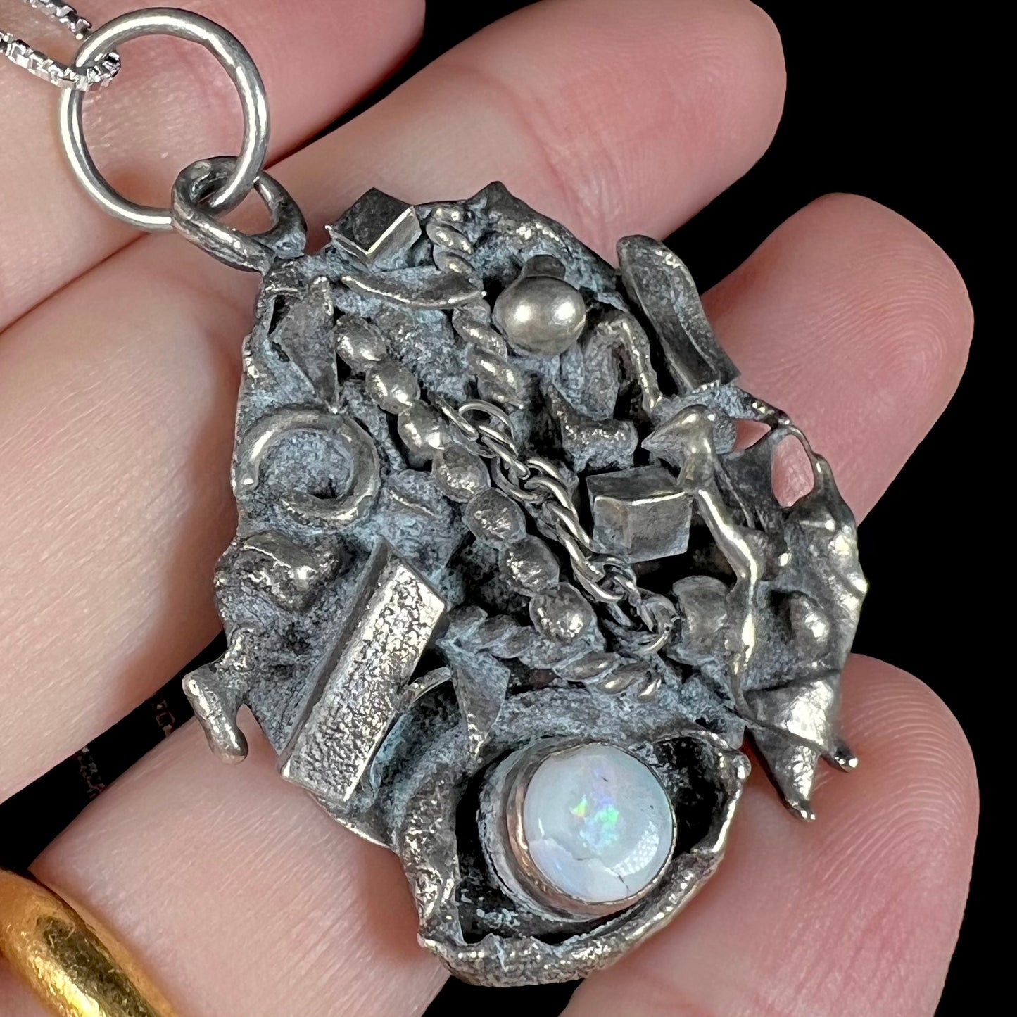 A brutalist scrap silver opal pendant depicting a shipwreck, shown close-up from a side angle.