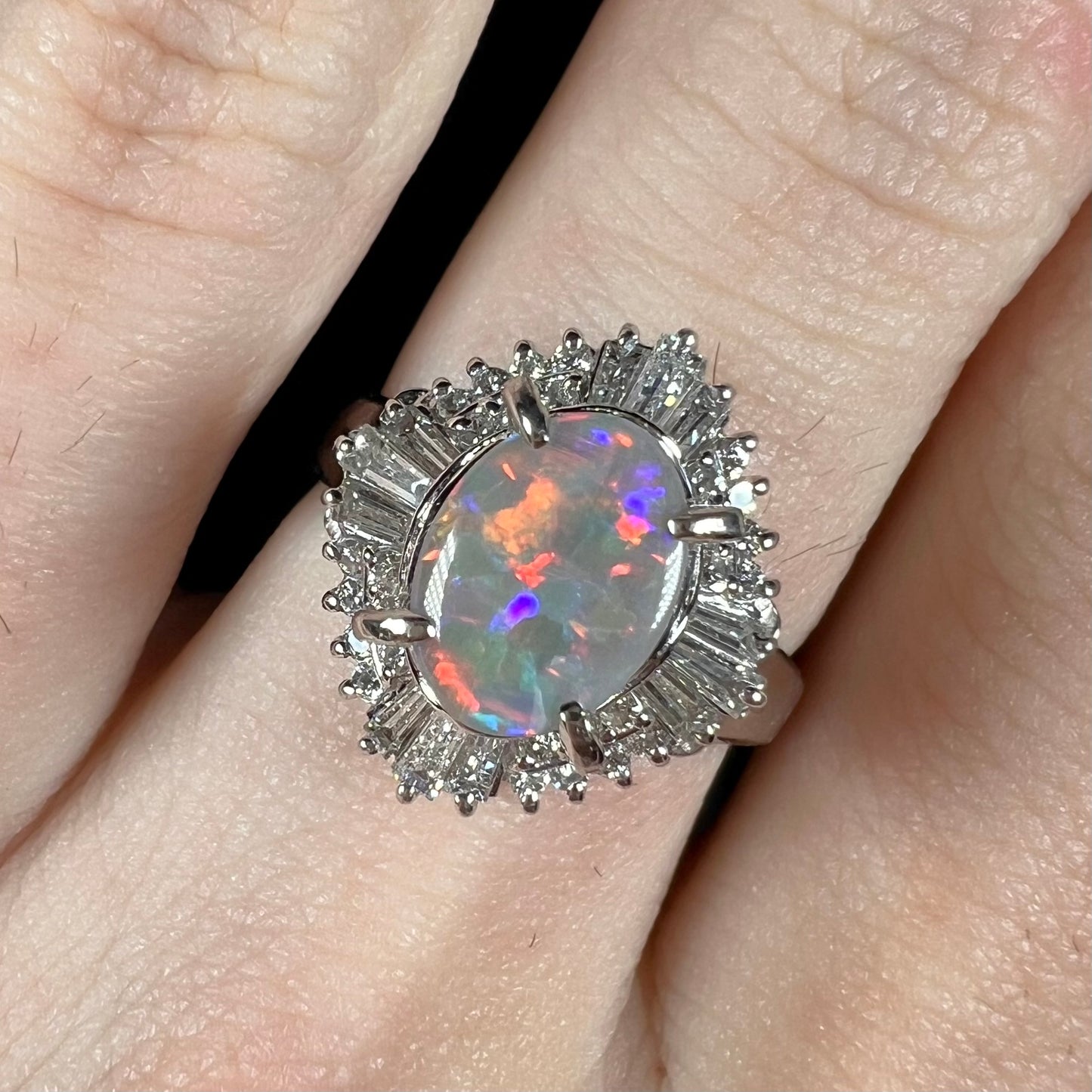 Close-up of a semi-black opal and diamond halo ring worn on a hand, highlighting its vintage platinum setting and red-blue opal fire.