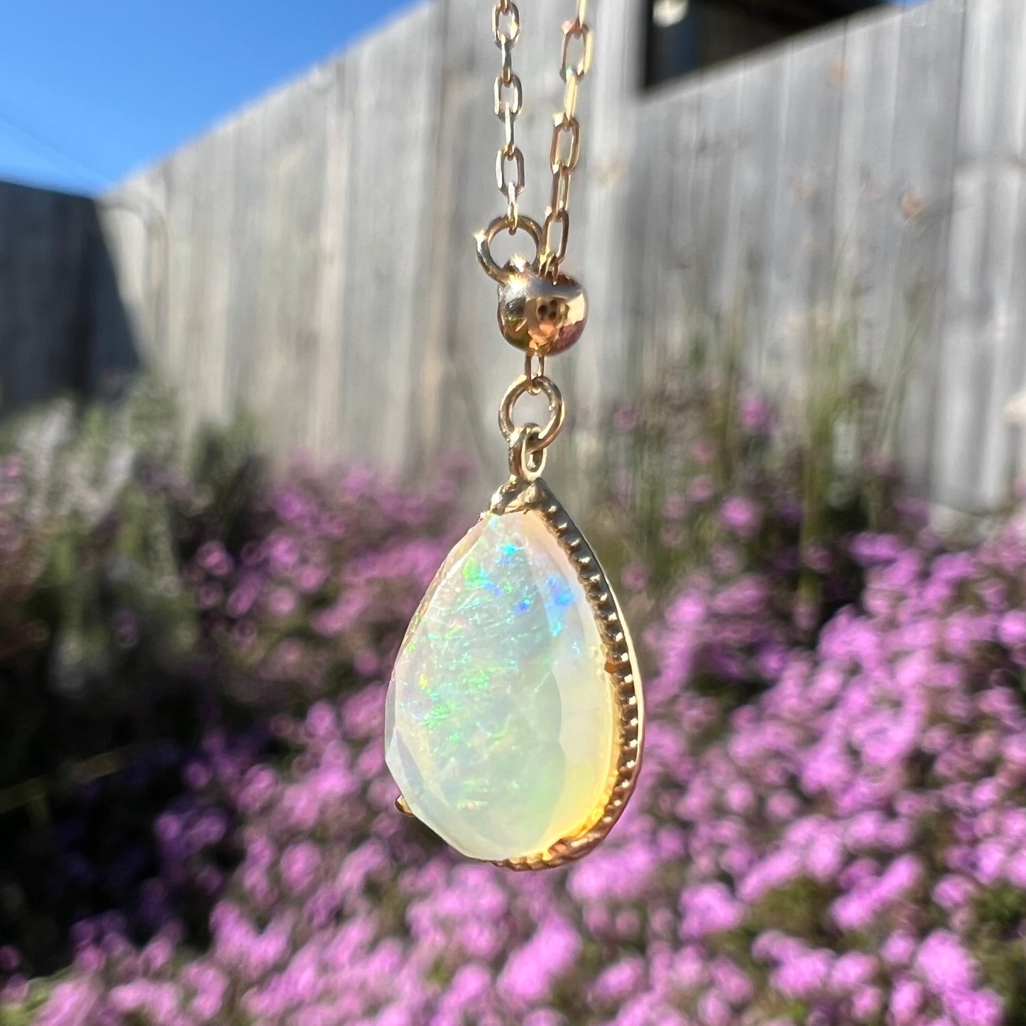 Side view of the opal necklace in sunlight, displaying faint green and blue flashes. Purple flowers on a bush in the background.