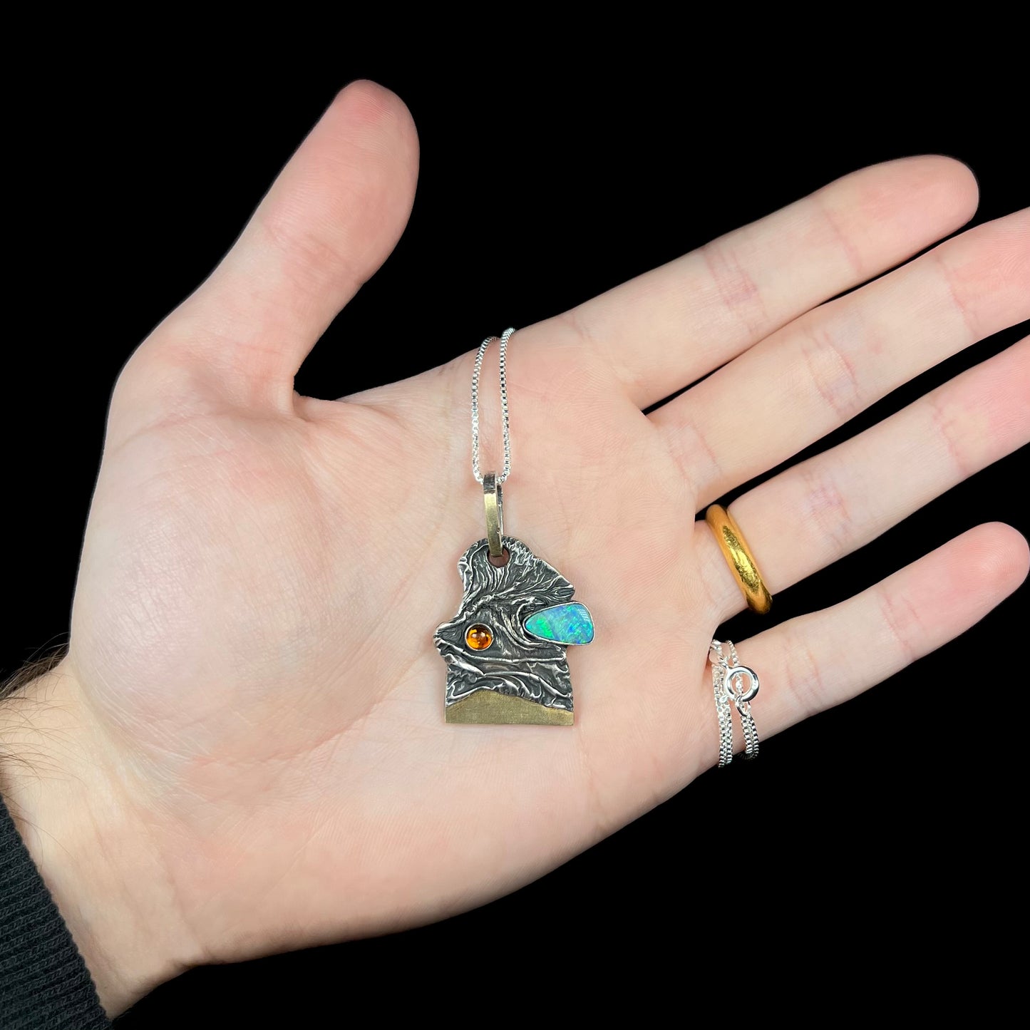 Hand holding a two-tone sterling silver and 18k gold-accented men's opal and amber necklace against a black background.