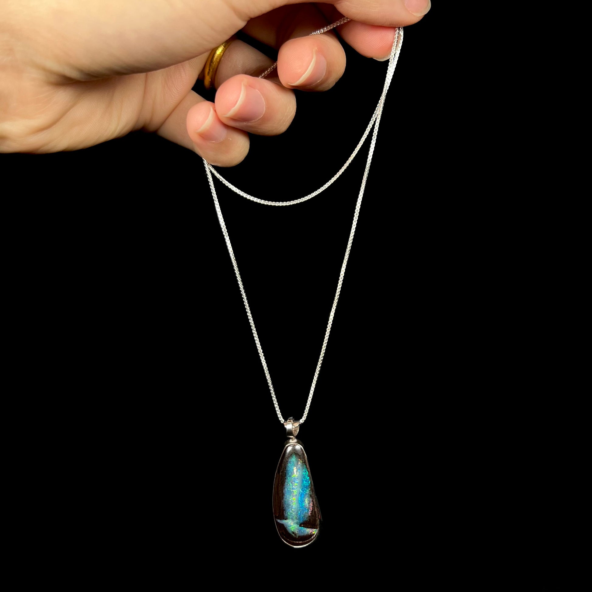 A sterling silver boulder opal necklace with blue color play, dangled from its chain by a hand.
