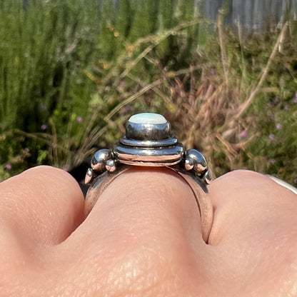 Close-up of a hand wearing a silver ring with a stone, set against a blurred natural background.