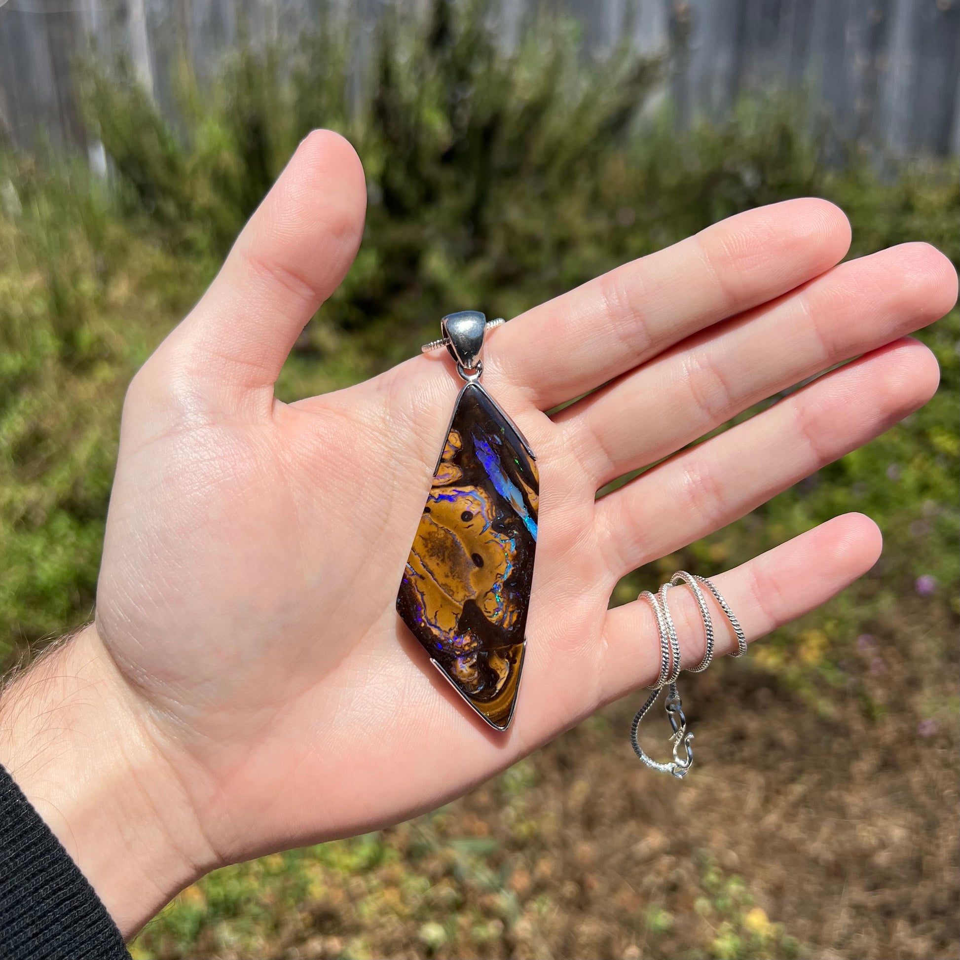 Hand holding large sterling silver Australian boulder opal men's pendant with freeform shape in sunlight