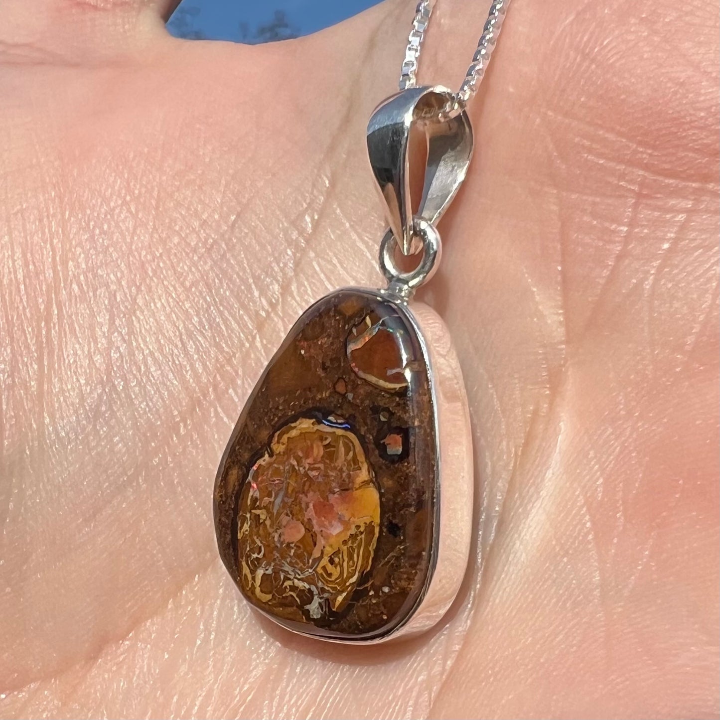 Close-up angled view of a Yowah nut boulder opal pendant in sterling silver, shown in the sunlight held in a hand.