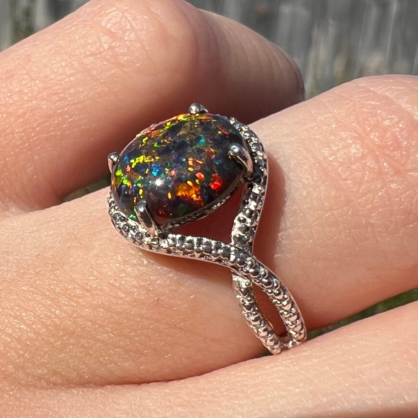 A lab-created black opal solitaire ring in sterling silver, shown at an angle on a finger in sunlight.