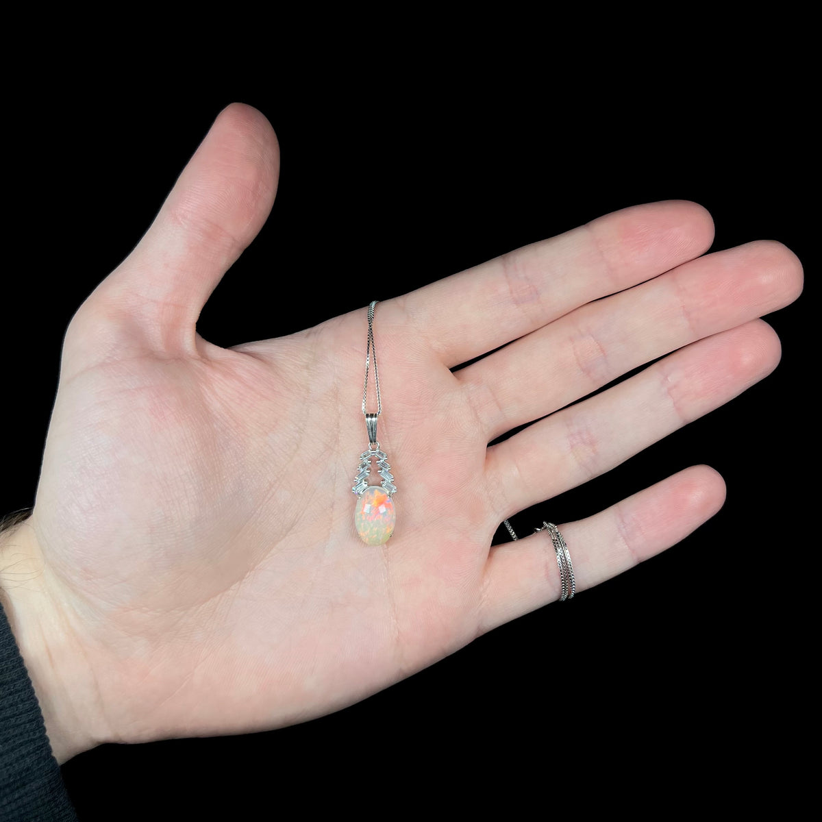 Hand holding silver Ethiopian opal and emerald cut CZ drop necklace; the chain is wrapped around pinky finger