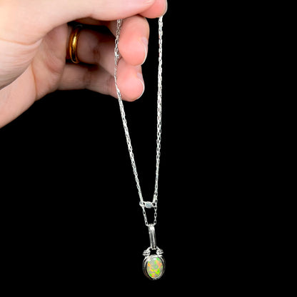 A colorful sterling silver Ethiopian opal necklace, dangled by its chain from a hand against a black backdrop.