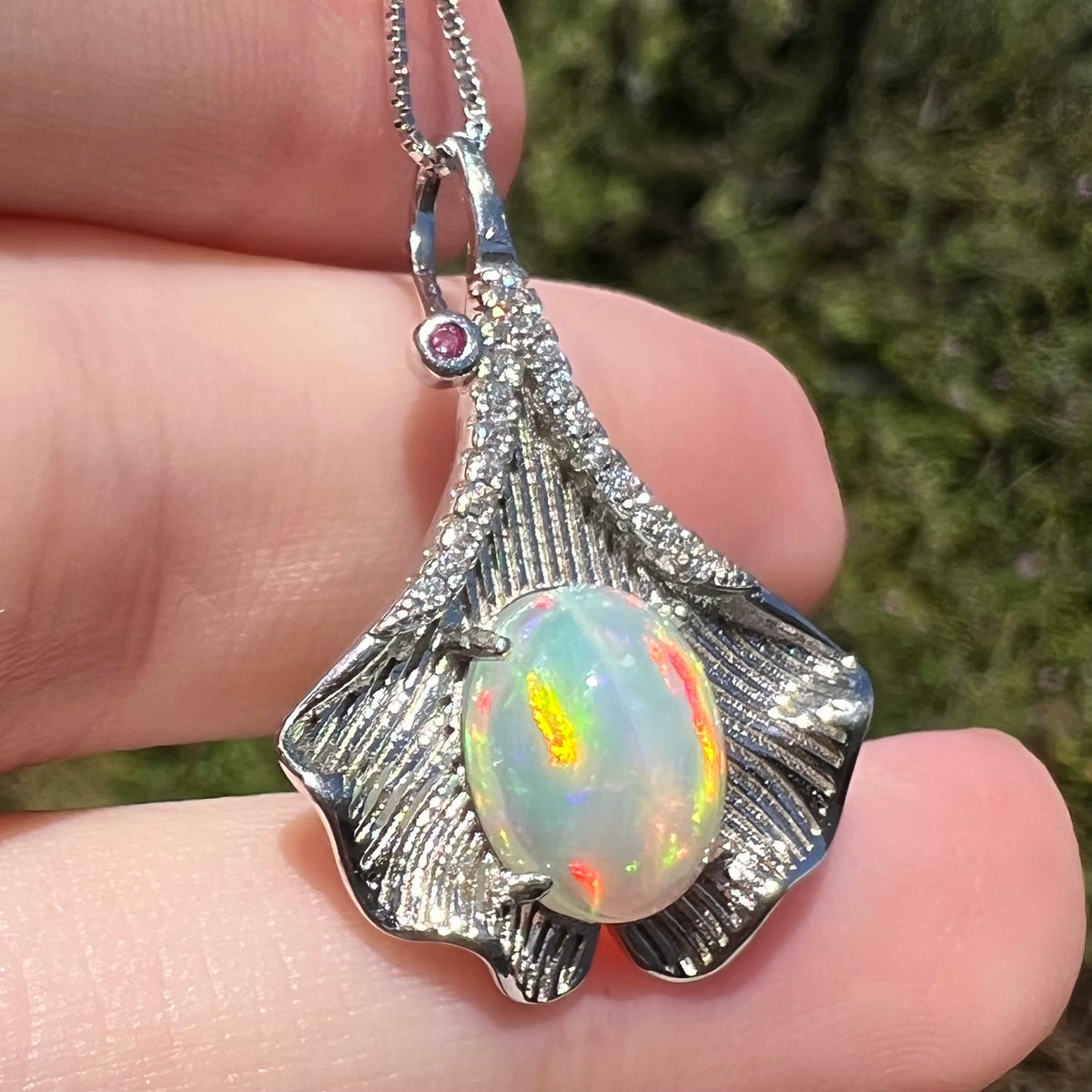 Sunlit close-up of sterling silver ginko leaf pendant set with red and orange Ethiopian opal, held in hand