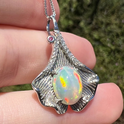 Sunlit close-up of sterling silver ginko leaf pendant set with red and orange Ethiopian opal, held in hand