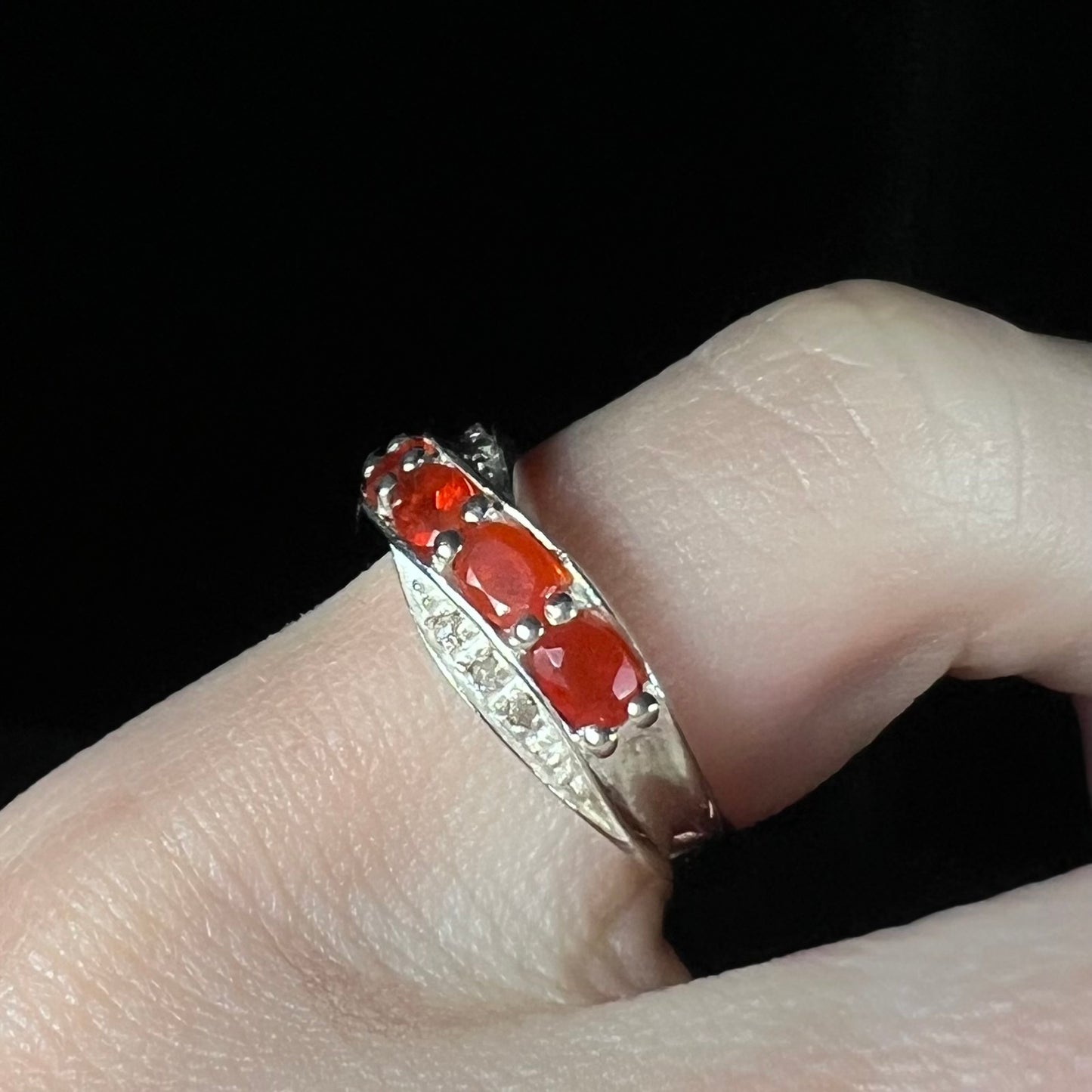 Angled close-up of silver ring with faceted fire opal gemstones worn on a finger against black background.