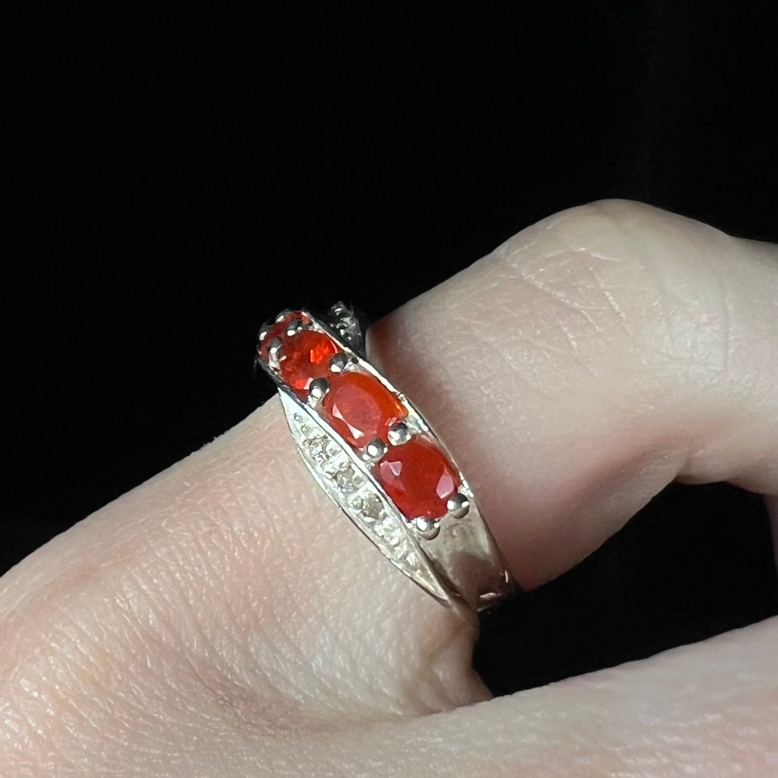 Angled close-up of silver ring with faceted fire opal gemstones worn on a finger against black background.