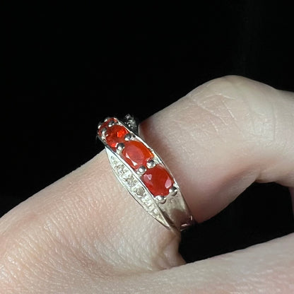 Angled close-up of silver ring with faceted fire opal gemstones worn on a finger against black background.