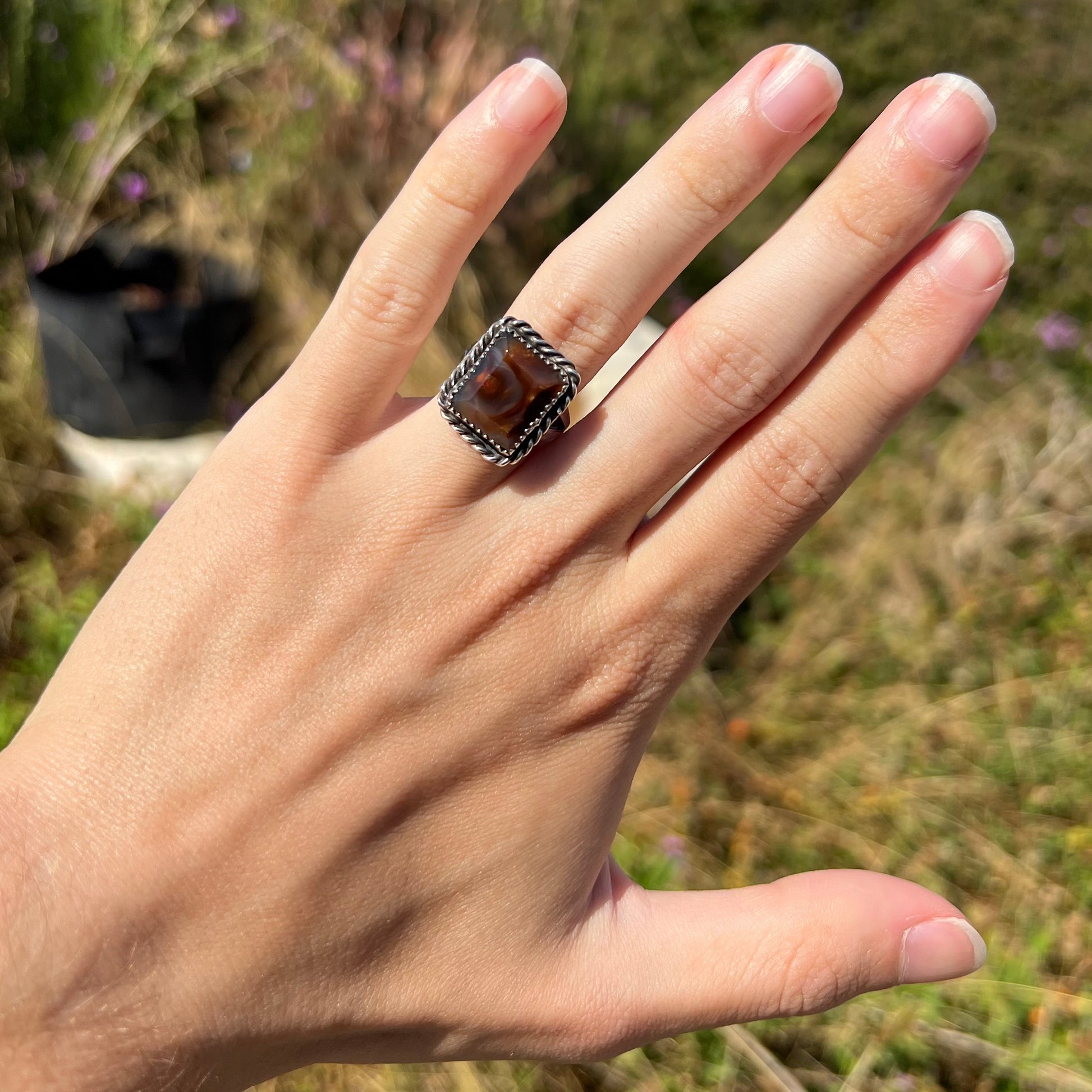 A sterling silver fire agate ring worn on a hand in the sunlight against blurred outdoor backdrop.