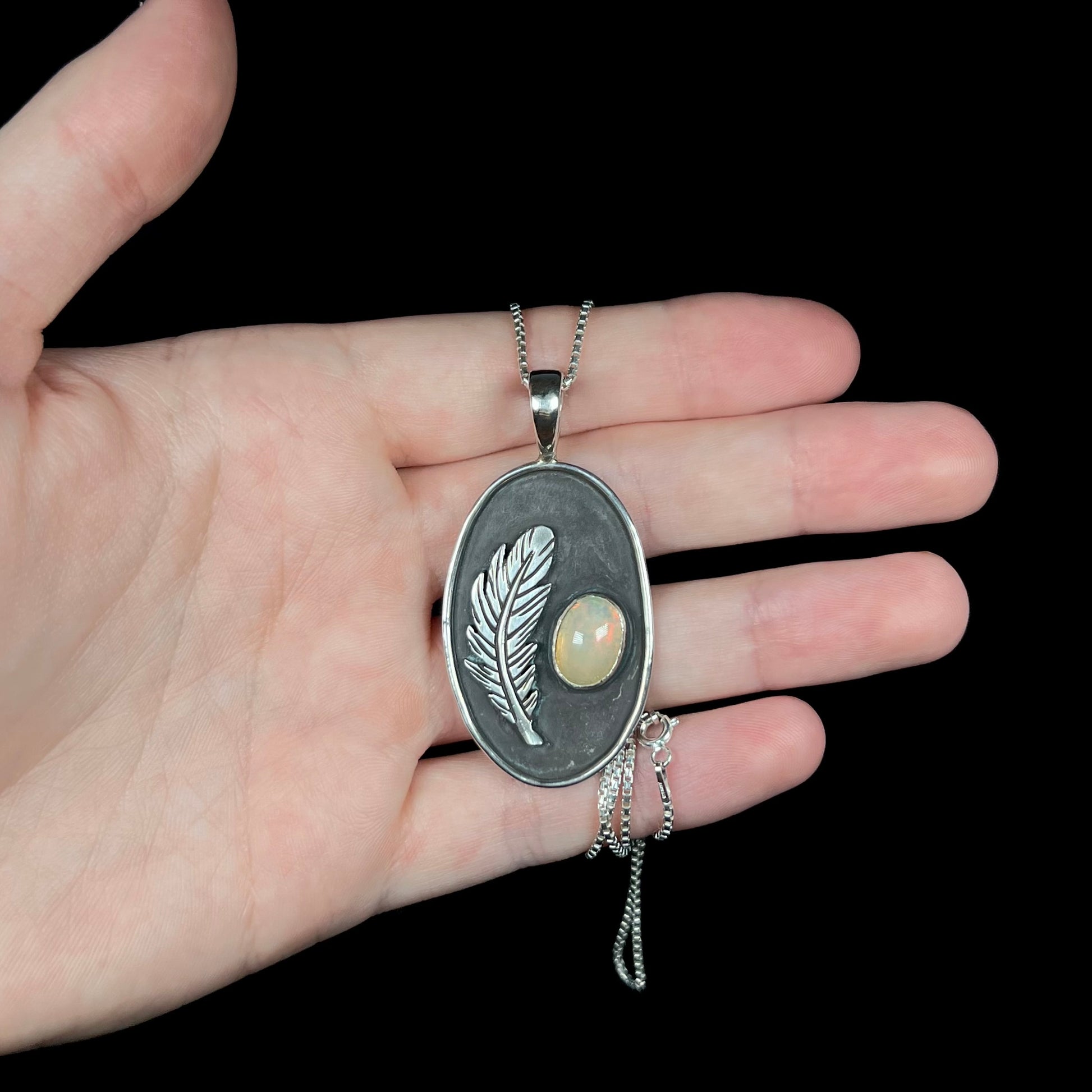 Hand holding a large sterling silver fire opal and feather design men's necklace under cool studio light against black backdrop.