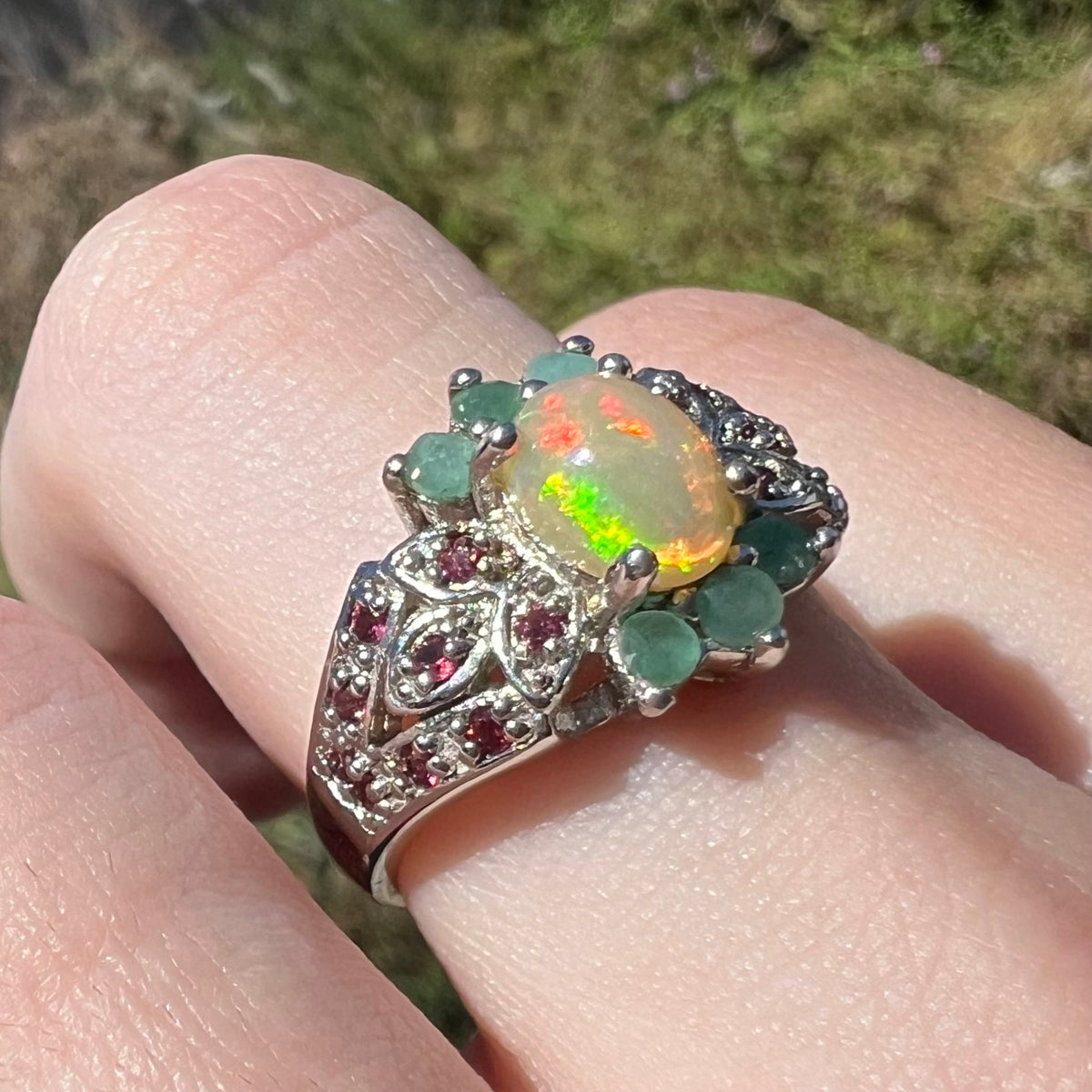 A silver ring with a large iridescent opal center stone and both emerald and garnet gemstone accents, shown worn on a person's finger with an outdoor blurred background.
