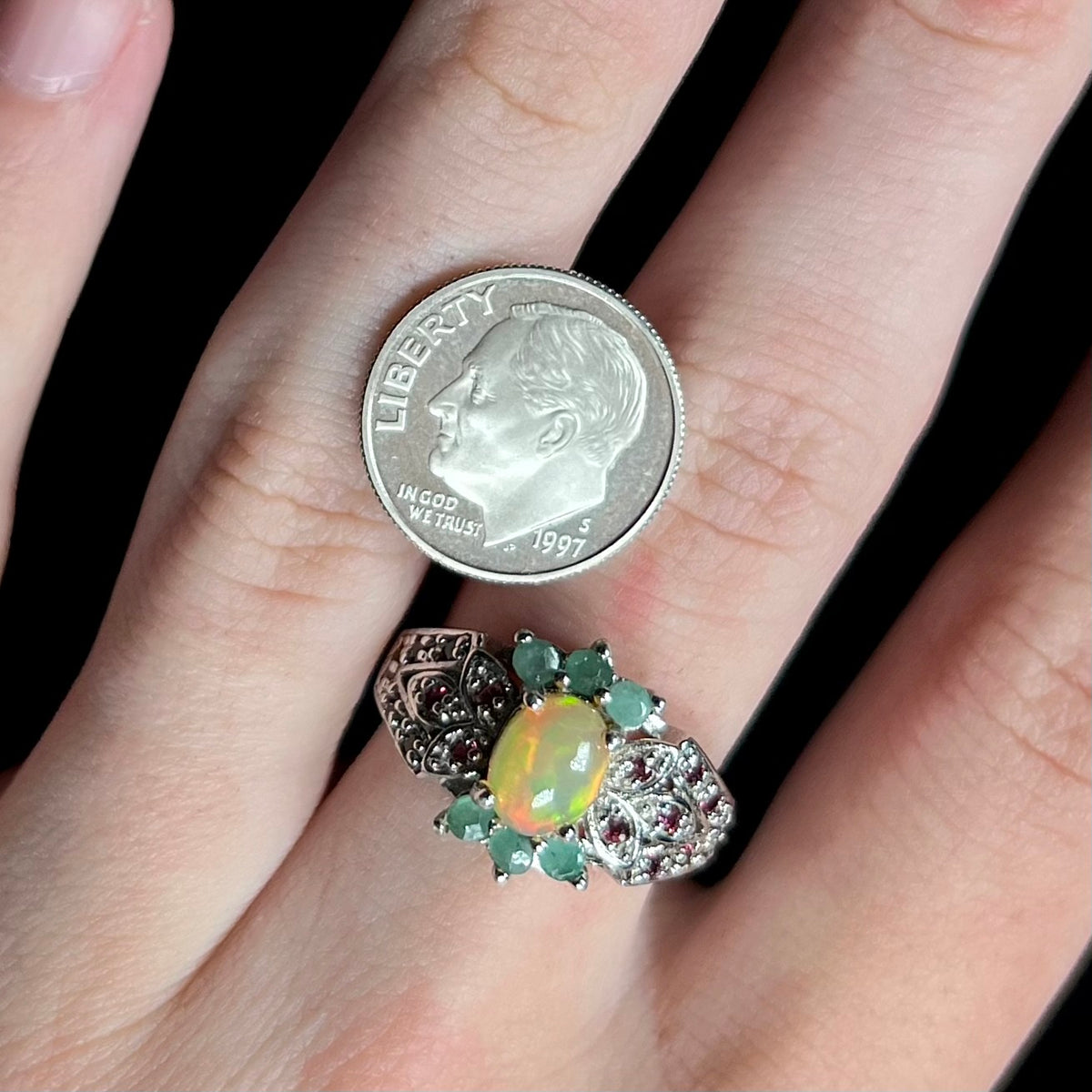 Close-up of hand wearing sterling silver Ethiopian fire opal, emerald, and garnet ring next to US dime coin