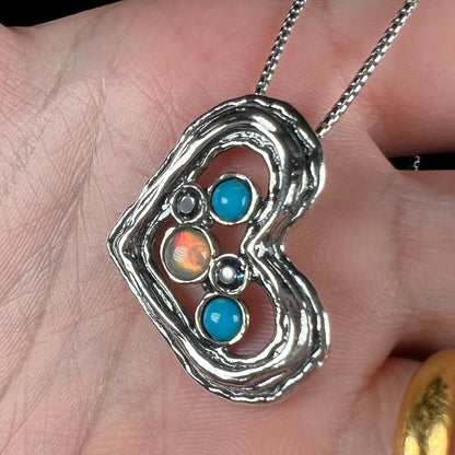 A sterling silver heart necklace set with opal, turquoise, and blue topaz shown close-up in a hand from an angle.